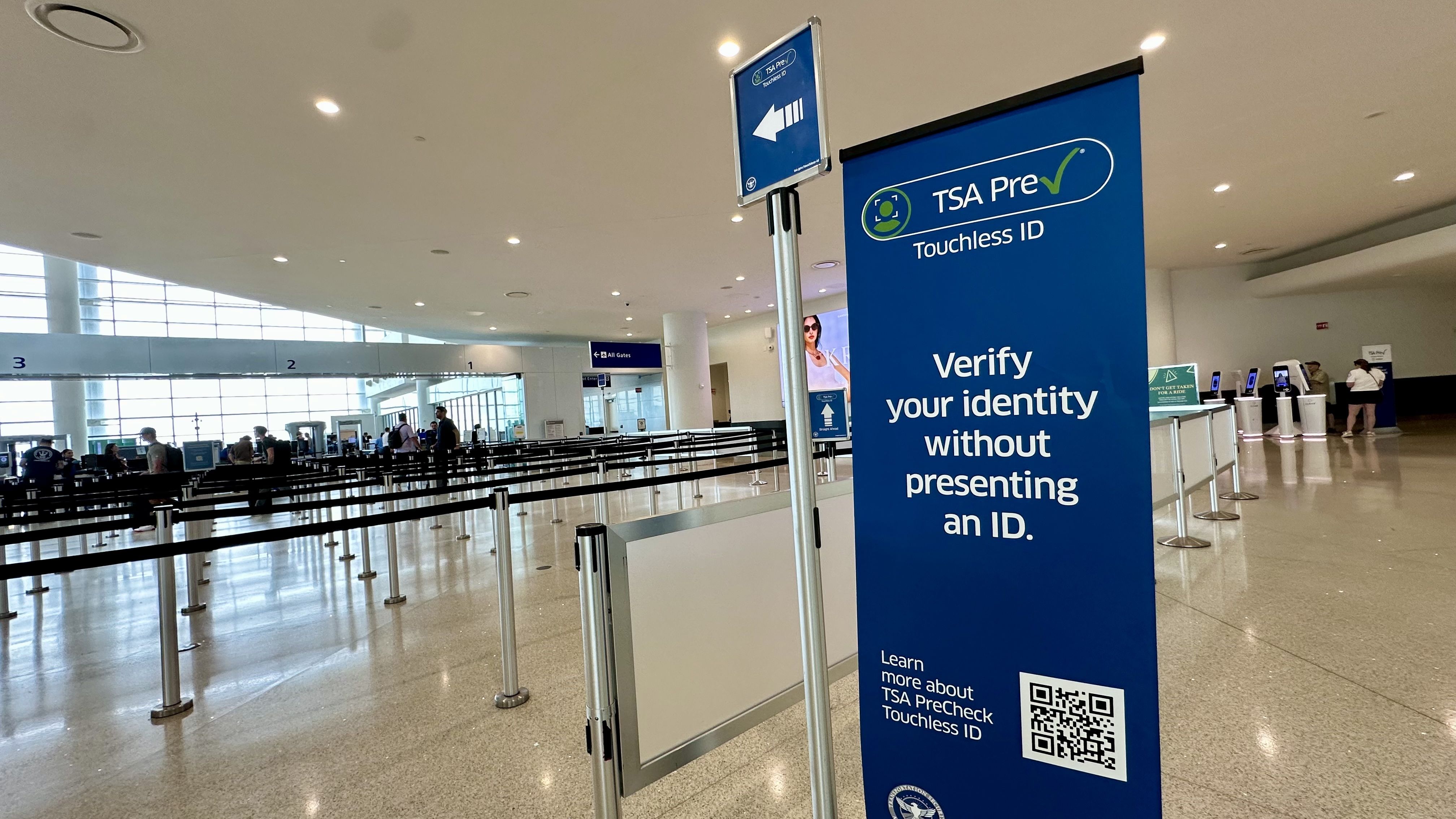Inside a bright airport, a large blue TSA PreCheck banner reads "Touchless ID" with "Verify your identity without presenting an ID." People queue at kiosks behind metal stanchions.