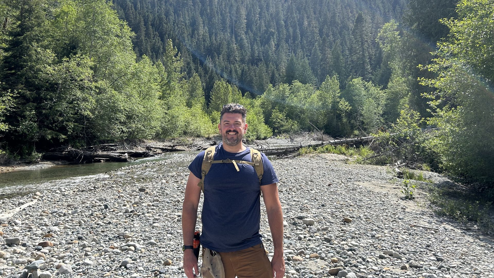 Man in a navy blue shirt and brown pants with a backpack stands on a rocky riverbank. Behind him are green trees and a mountain under a partly cloudy blue sky.