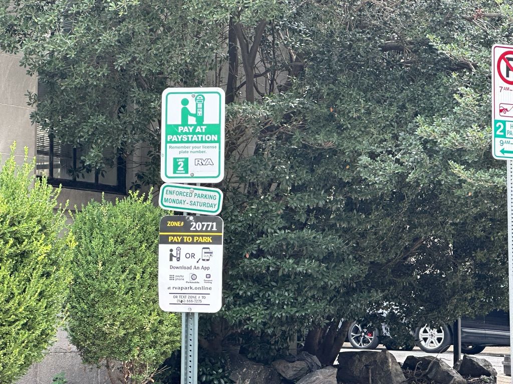 A downtown sign stack tells drivers to pay at a kiosk, while a newer placard on the same pole tells them to use an app or text a zone number.