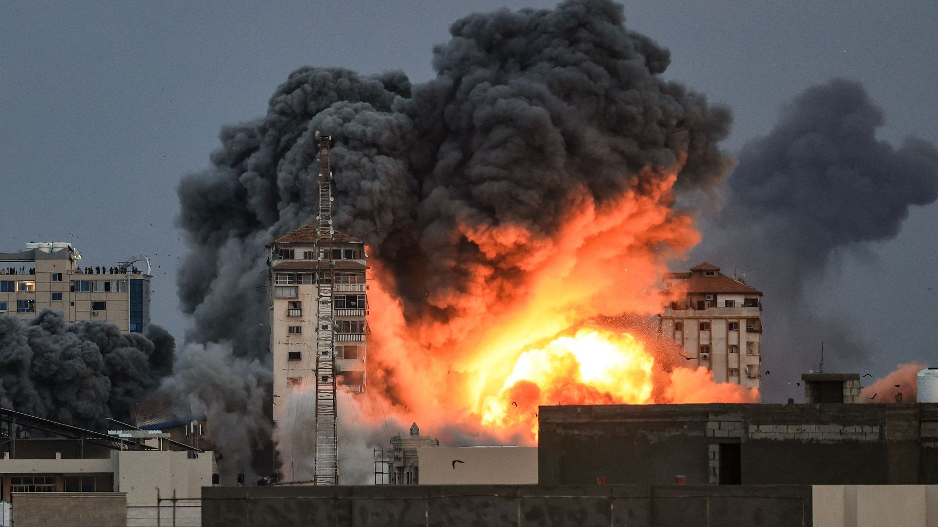 People standing on a rooftop watch as a ball of fire and smoke rises above a building in Gaza City on October 7, 2023 during an Israeli air strike