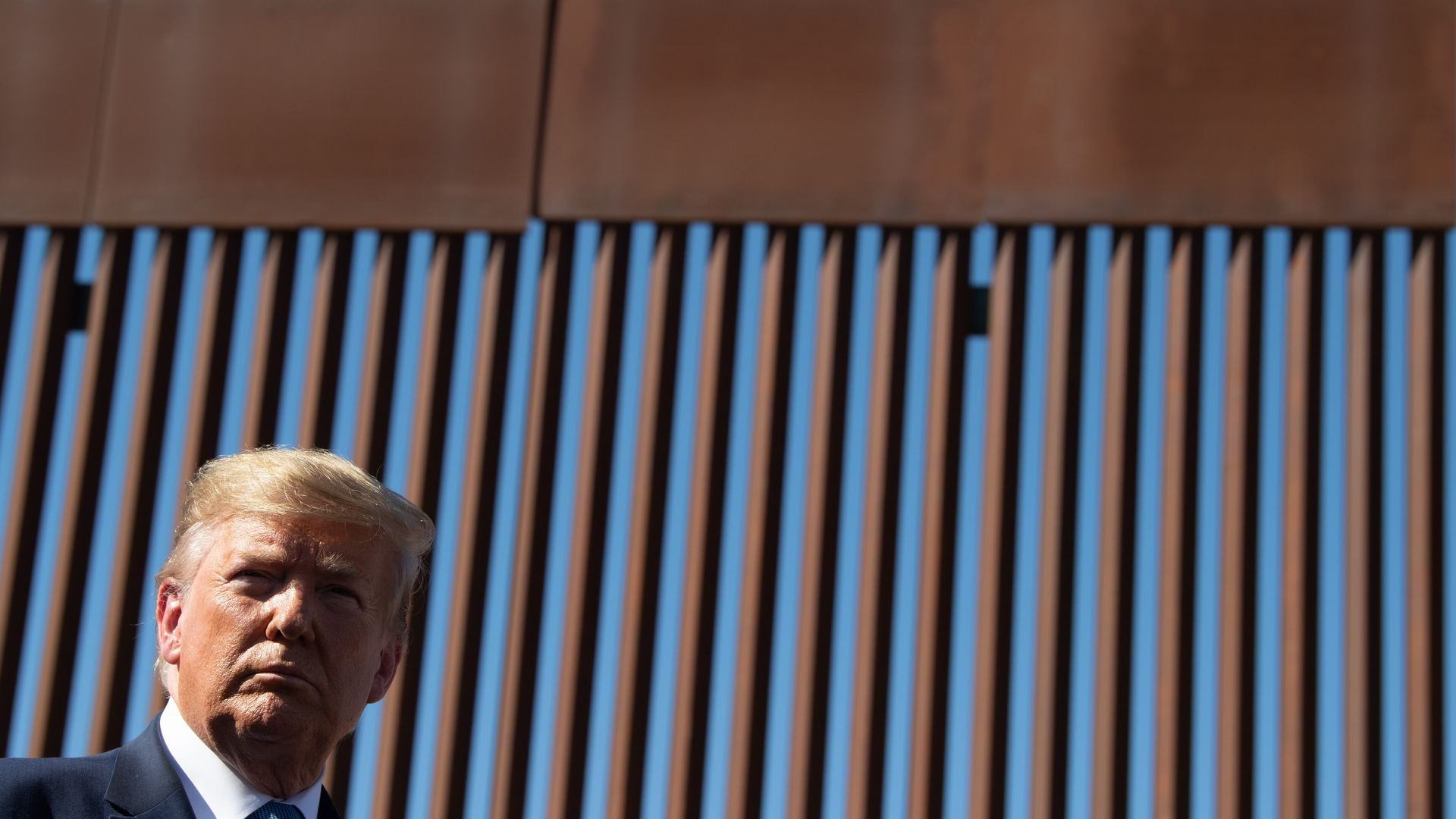 Donald Trump's head in the bottom left corner with the border wall — with metal slats — in the background. 