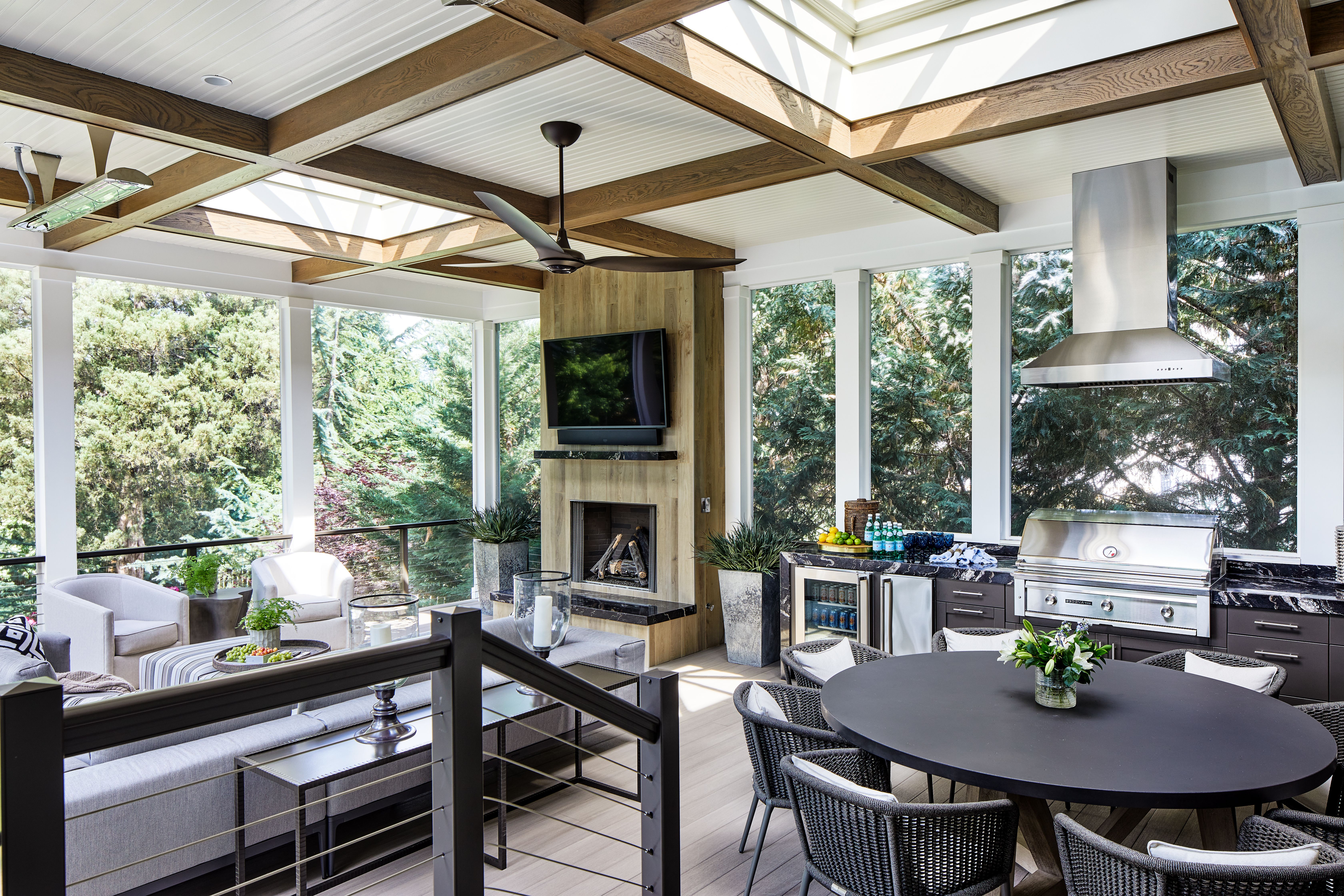 Bright screened porch with exposed wood beams, skylights, and a ceiling fan. A fireplace with a mounted TV, lounge seating, a round black dining table, and a stainless outdoor kitchen with grill.