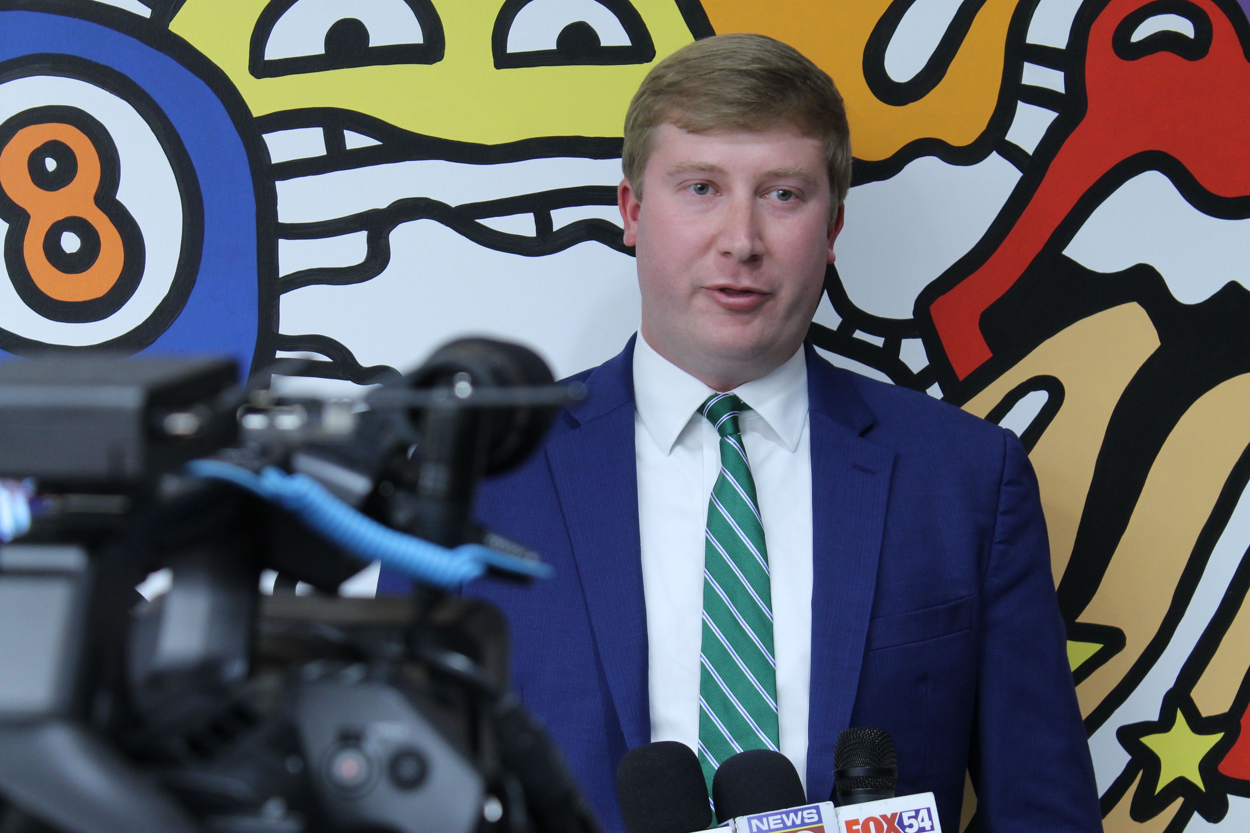 Portrait of a man in a blue suit and green striped tie speaking to reporters, with a colorful mural backdrop and several microphones in the foreground.