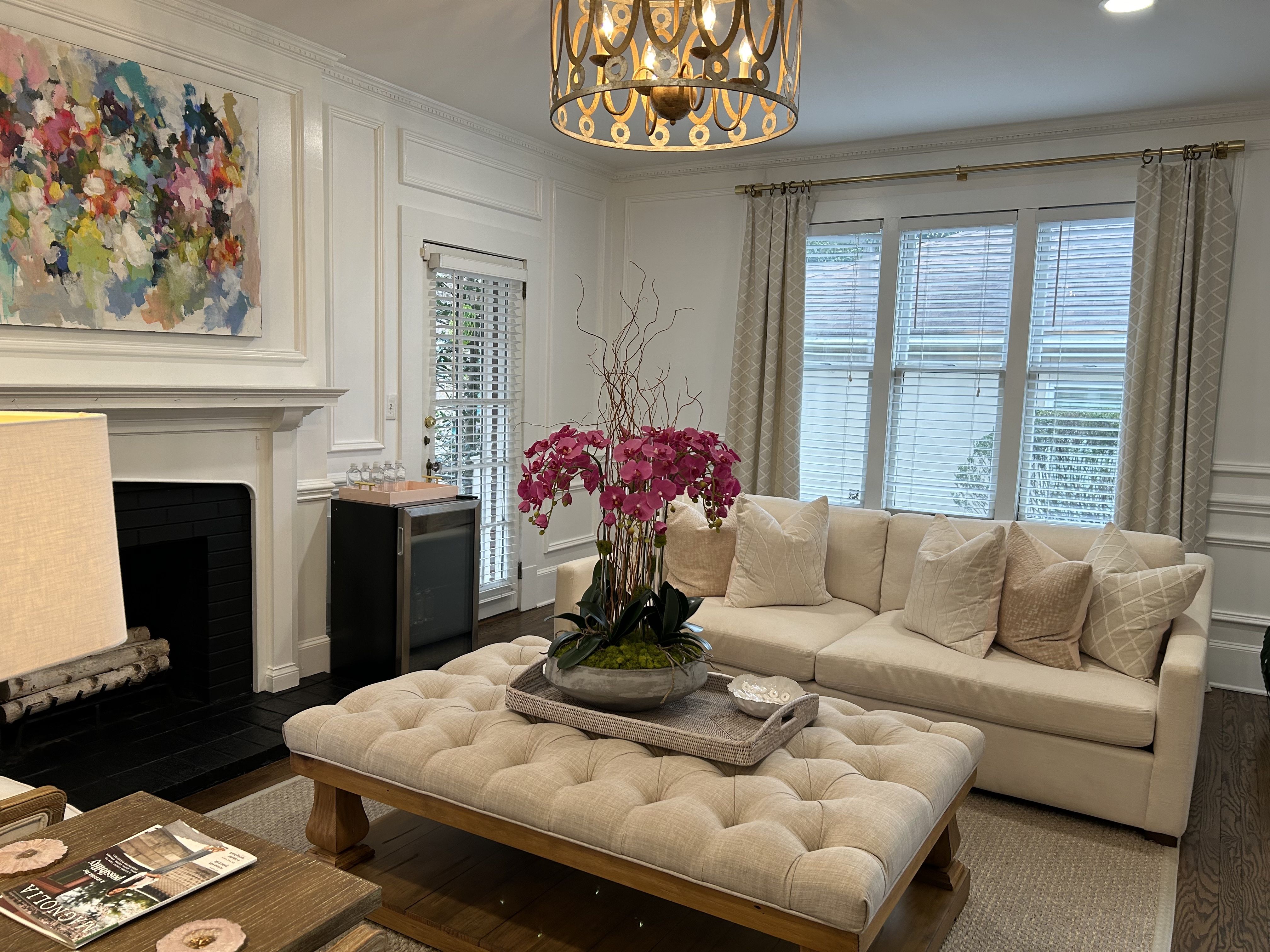 Cozy living room with beige sofa and cushions, tufted ottoman with pink orchid centerpiece, black fireplace, colorful abstract painting, large windows with blinds, and a decorative hanging light.