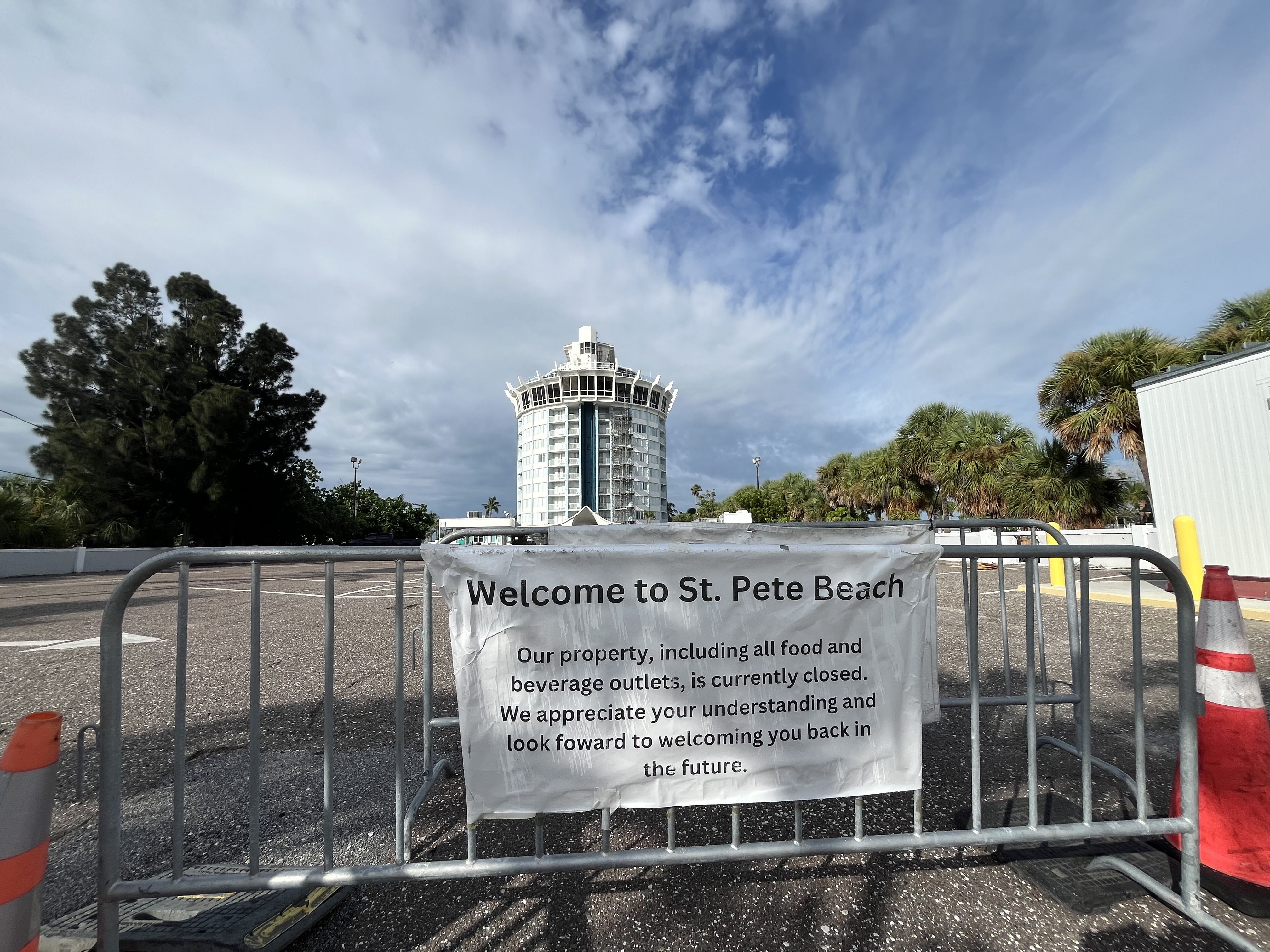 Metal barricade with a white sign reading "Welcome to St. Pete Beach. Our property, including all food and beverage outlets, is currently closed." A round, white building and palm trees are visible behind.