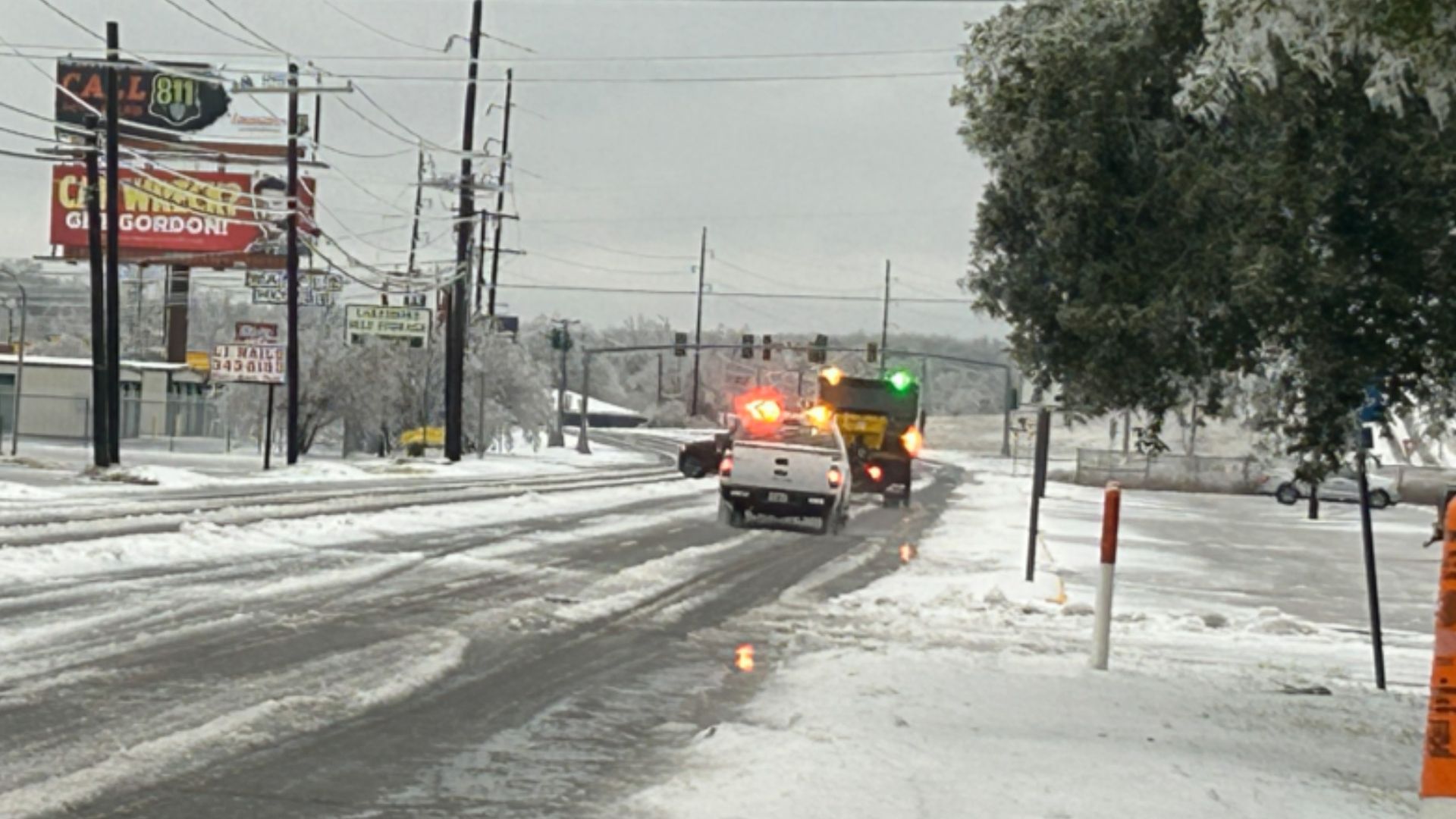 Snow-covered road with a white truck and snow plow clearing ice, street signs and poles on left, trees dusted with snow and orange traffic cone on right under gray sky.