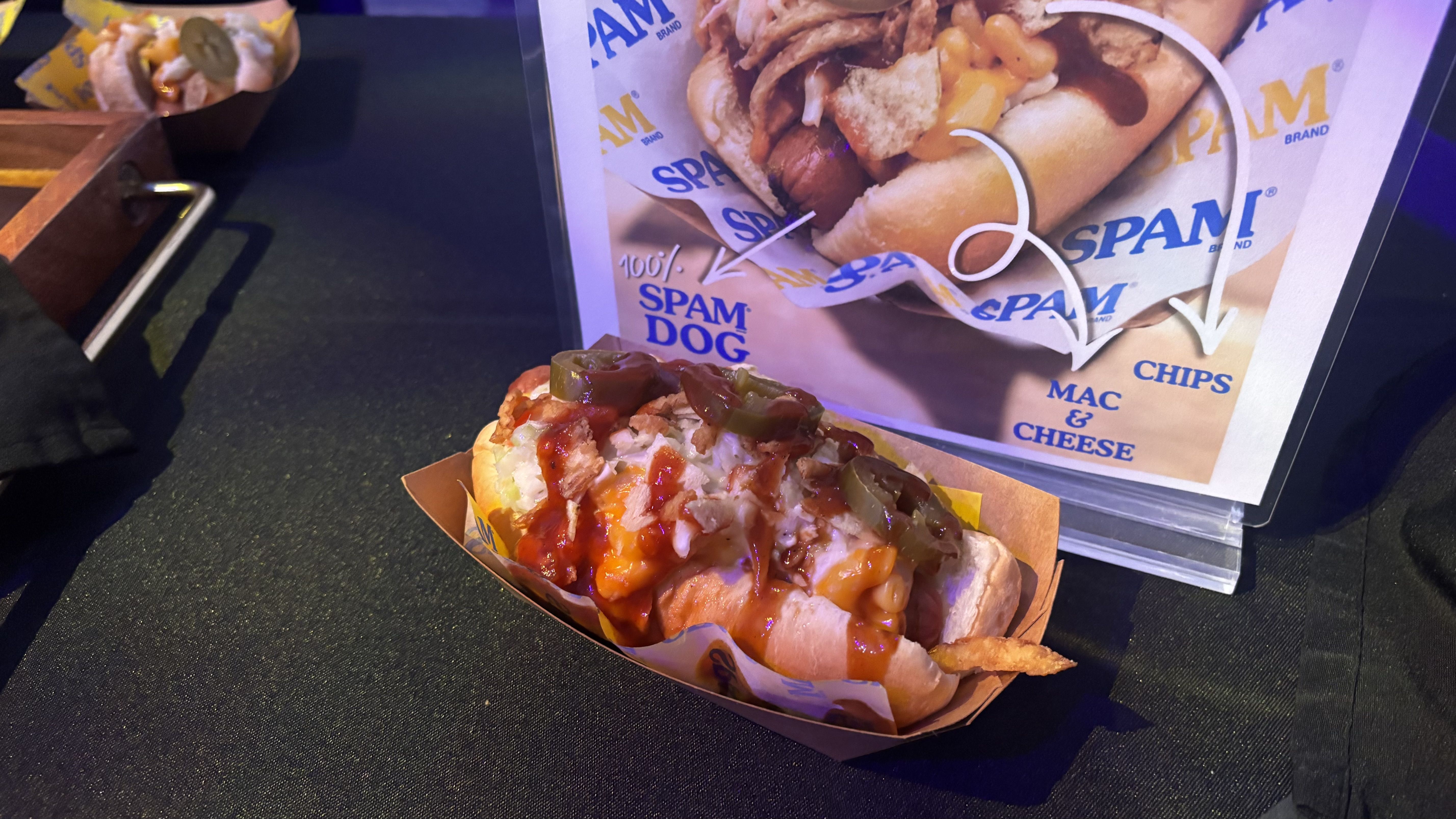 Two hot dogs in paper trays on a dark table; the front dog is topped with cheese, sauce, and jalapeños. A SPAM-themed menu poster with blue and yellow graphics is in the background.