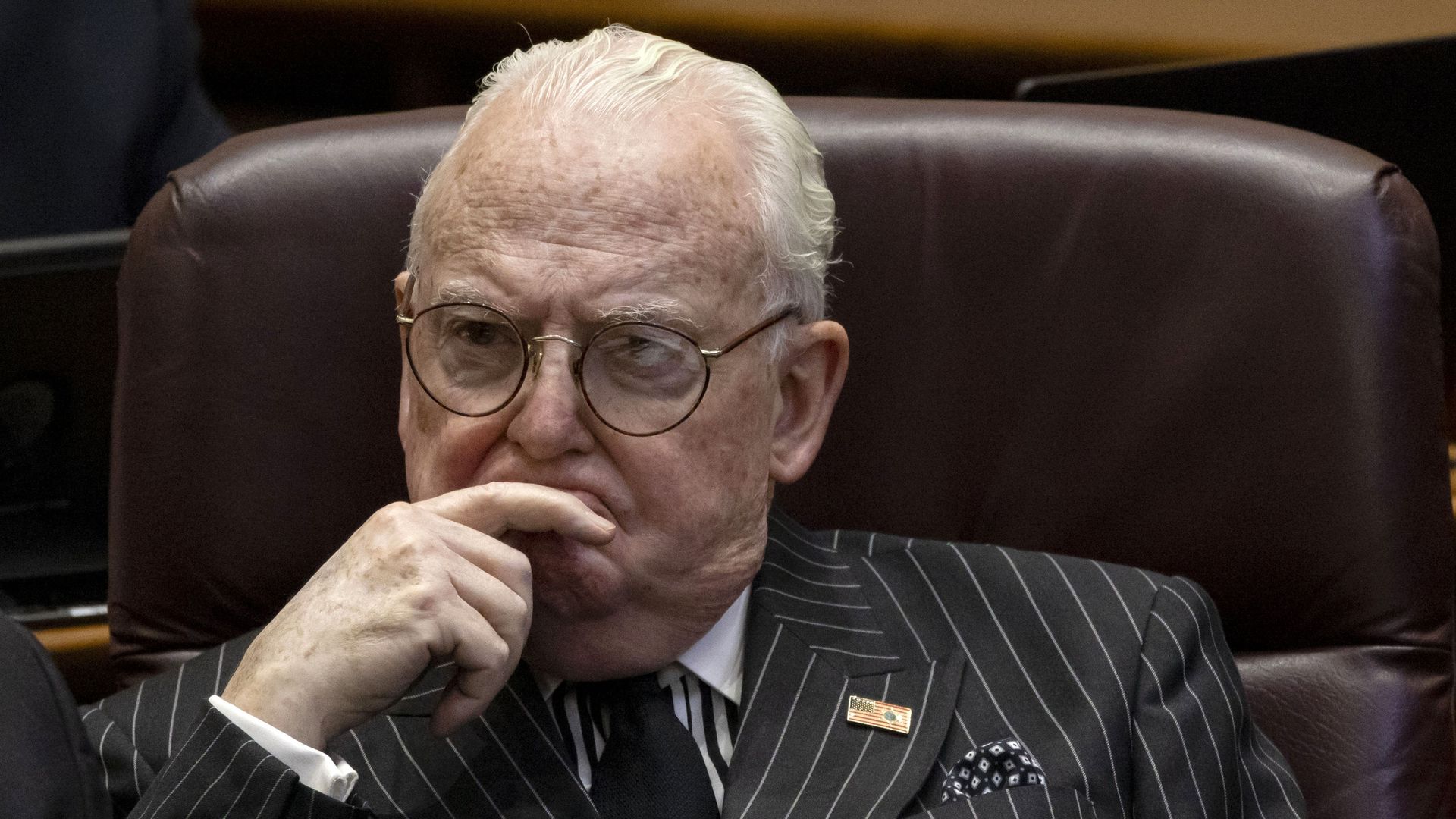 Man with hand up to his mouth, covering it, wearing glasses and pinstriped suit.