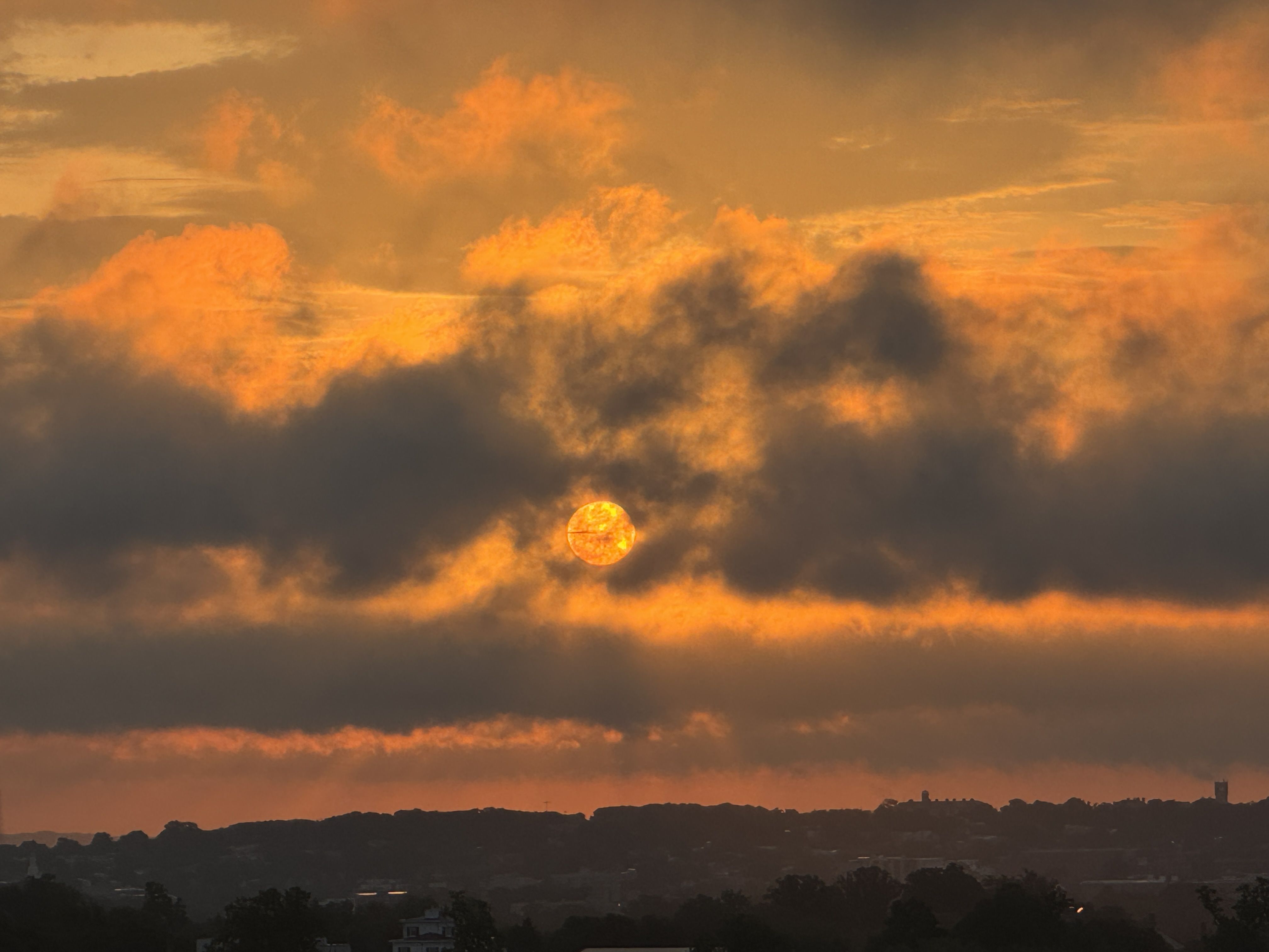 Sunset with bright orange clouds partially covering the sun above a dark silhouette of a town with trees and buildings.