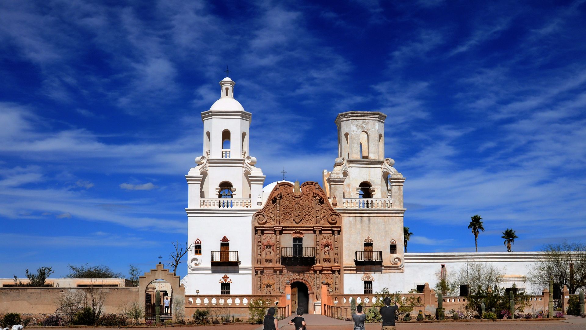 A Spanish colonial-style mission two towers on either side of the front entrance. A wall extends around the larger site in a desert landscape. 