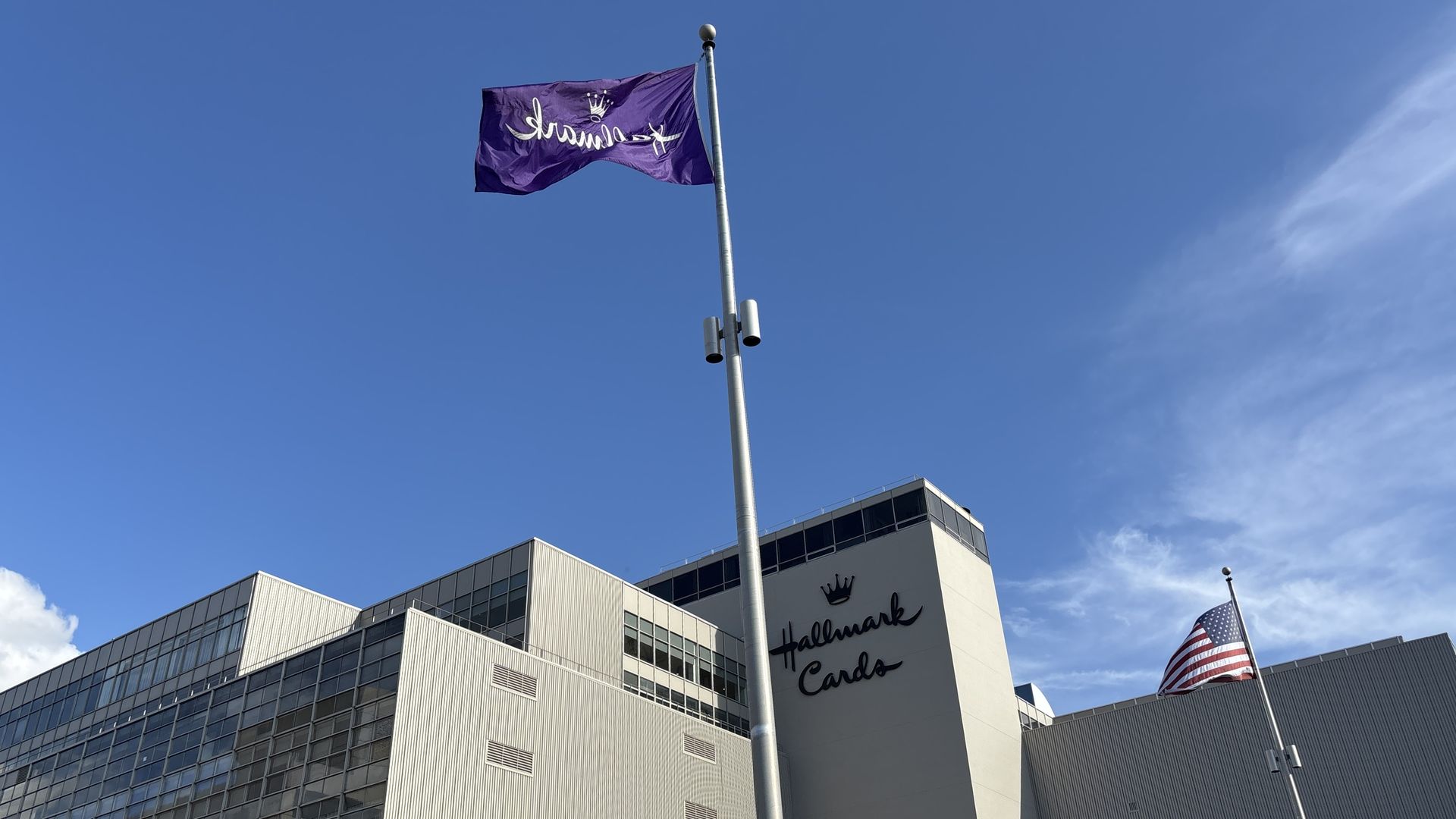 Purple flag with white cursive text and a crown emblem flutters on a tall flagpole above a modern building displaying Hallmark Cards; an American flag is visible to the right.