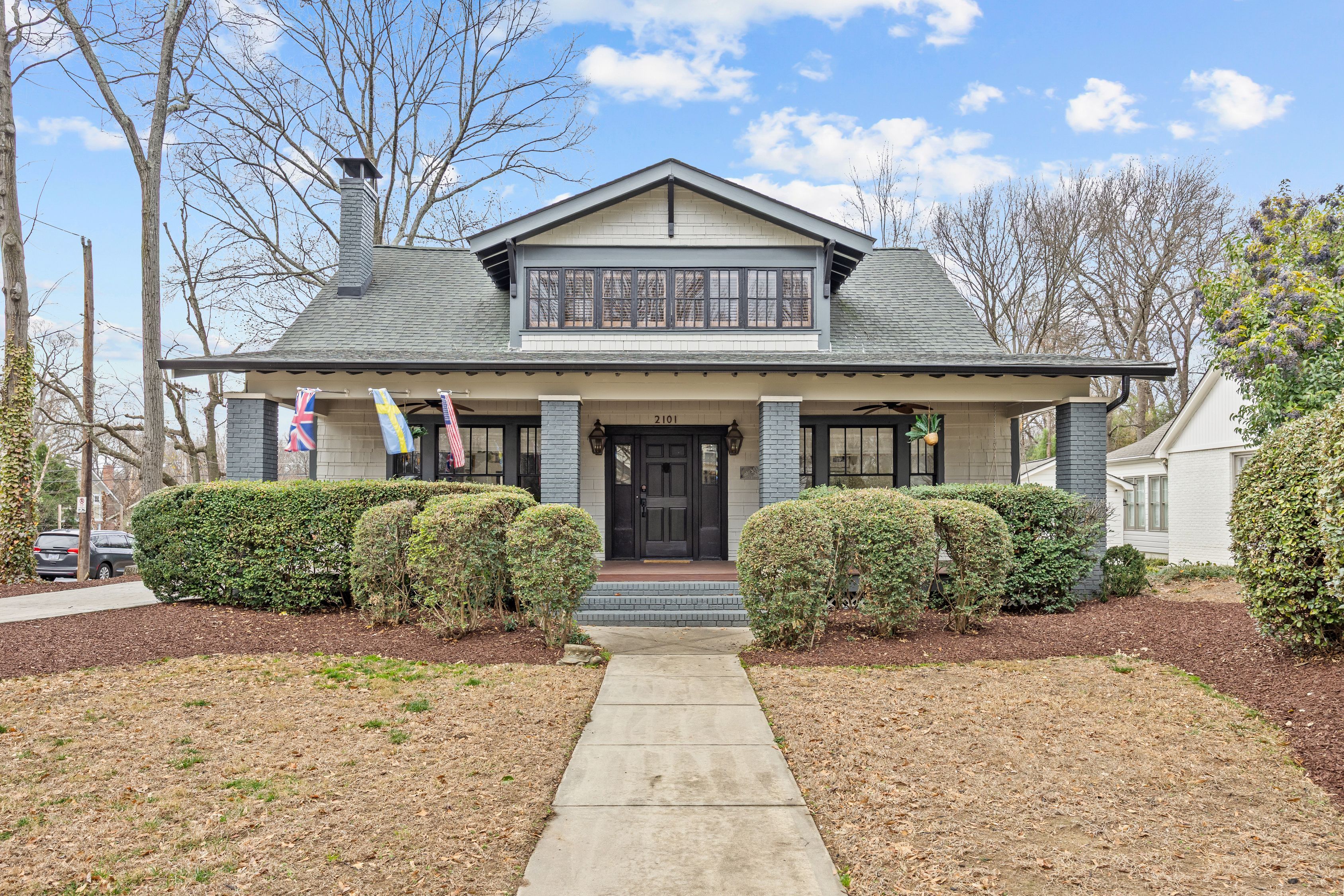 Gray and white two-story house with black door, front porch with three flags, rounded green bushes, stone walkway, and leafless trees under blue sky with clouds.