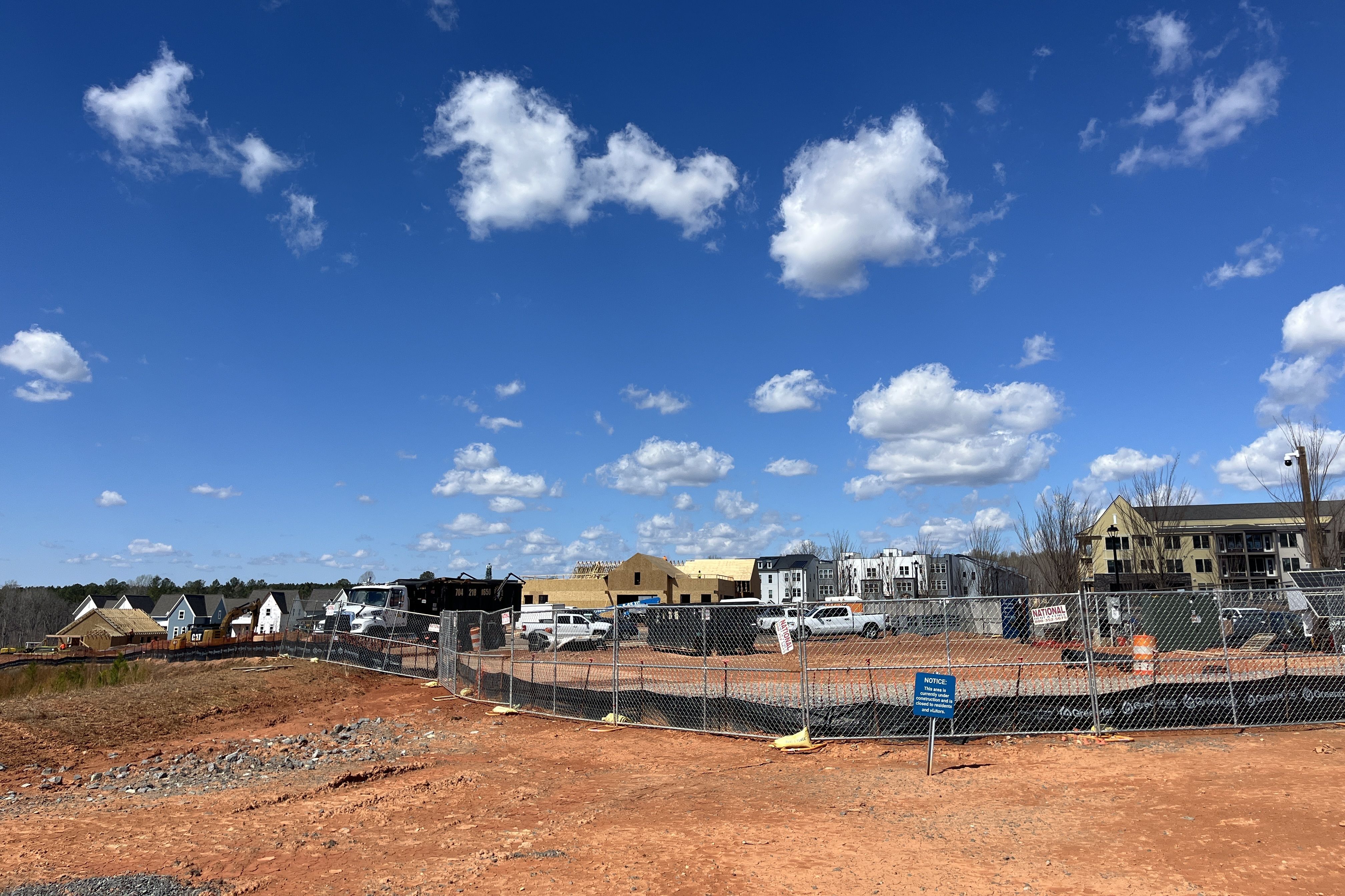 A construction site with orange dirt, chain-link fences, and several partially built houses in the background, with trucks nearby under a bright blue sky and white clouds.