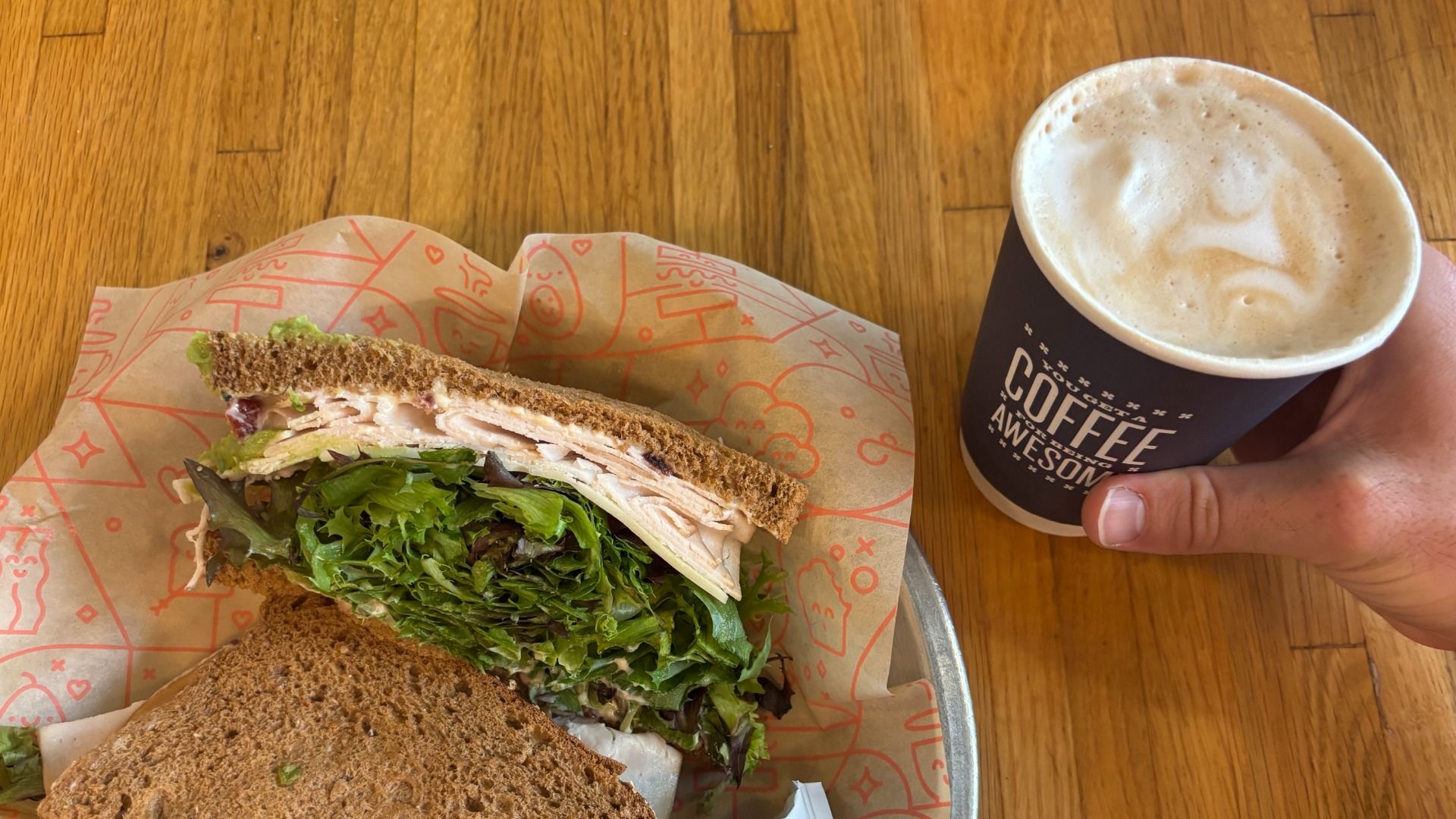 Top-down view of a hand holding a coffee cup with frothy latte beside a turkey and greens sandwich on brown bread, resting on a wooden table with orange-patterned parchment wrap.