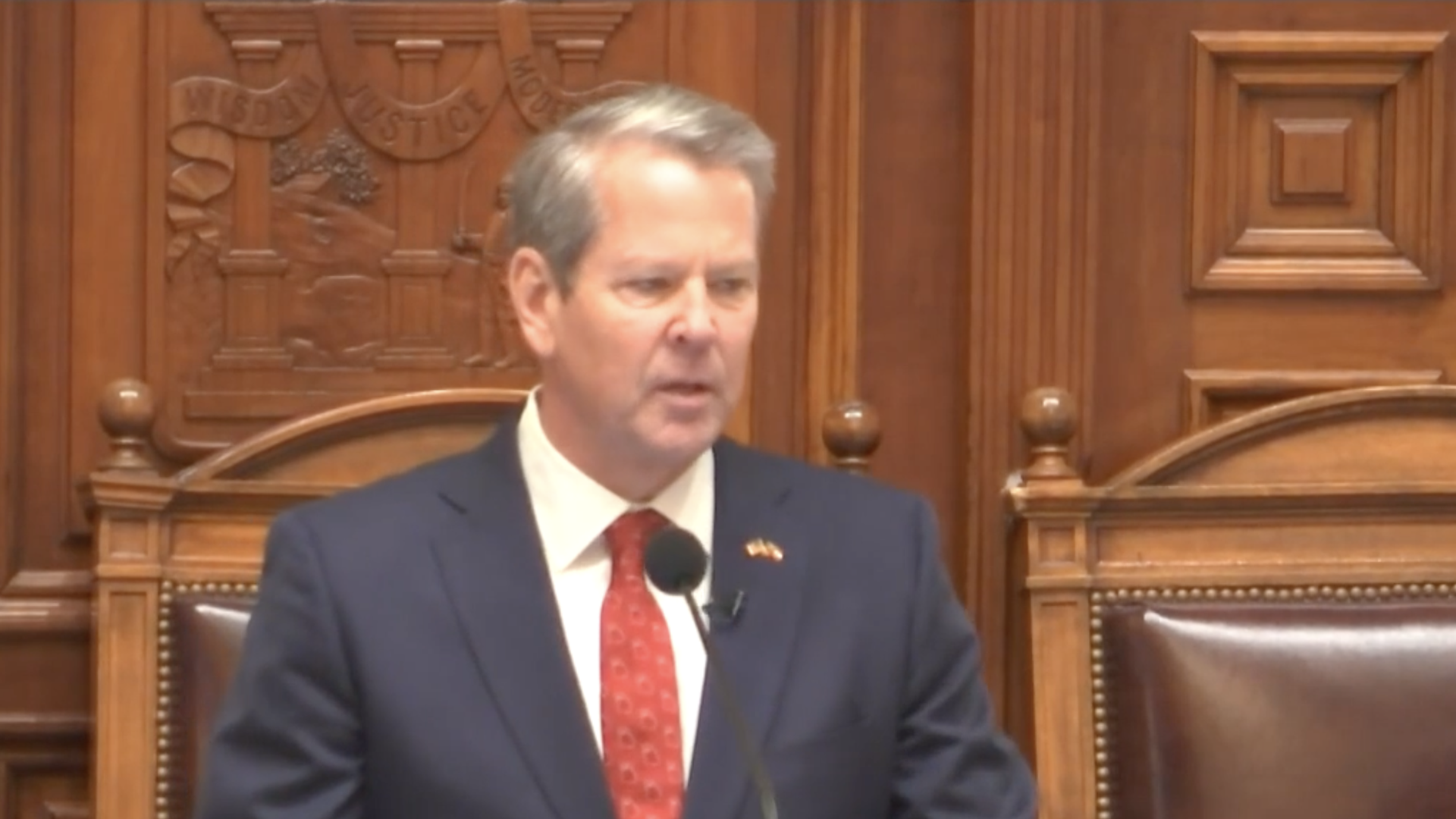 A screenshot of Gov. Brian Kemp speaking at a lectern in a grand legislative hall with carved wooden walls