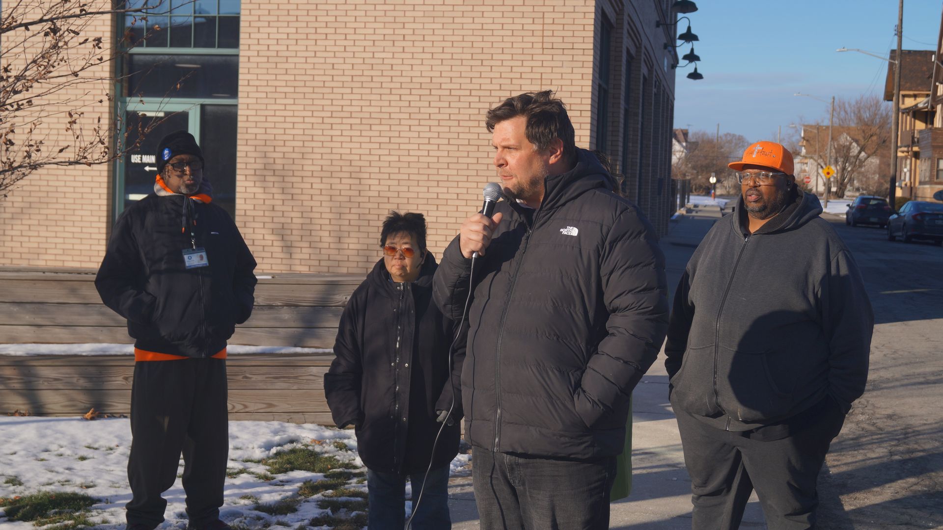 Man in black coat speaks at mic outdoors