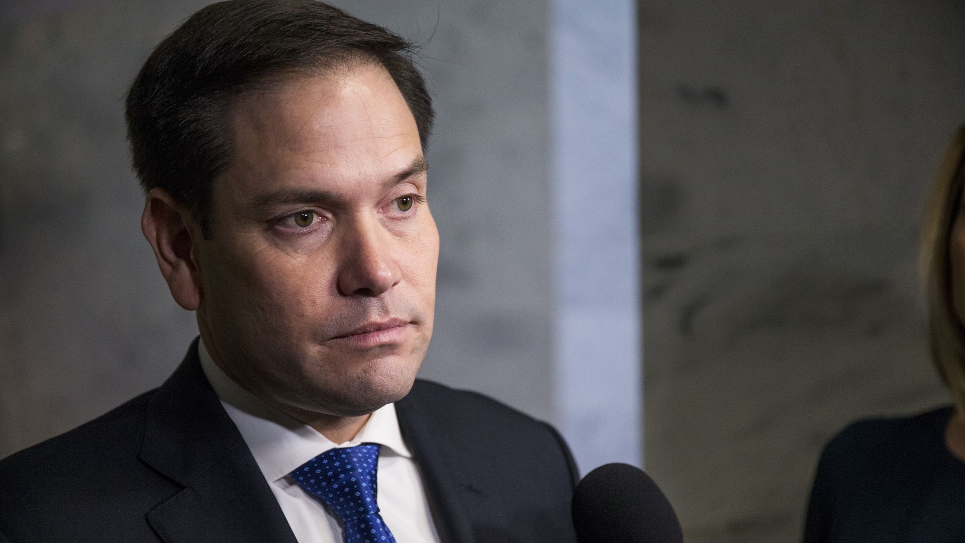 In this image, Marco Rubio looks ahead while facing a microphone.
