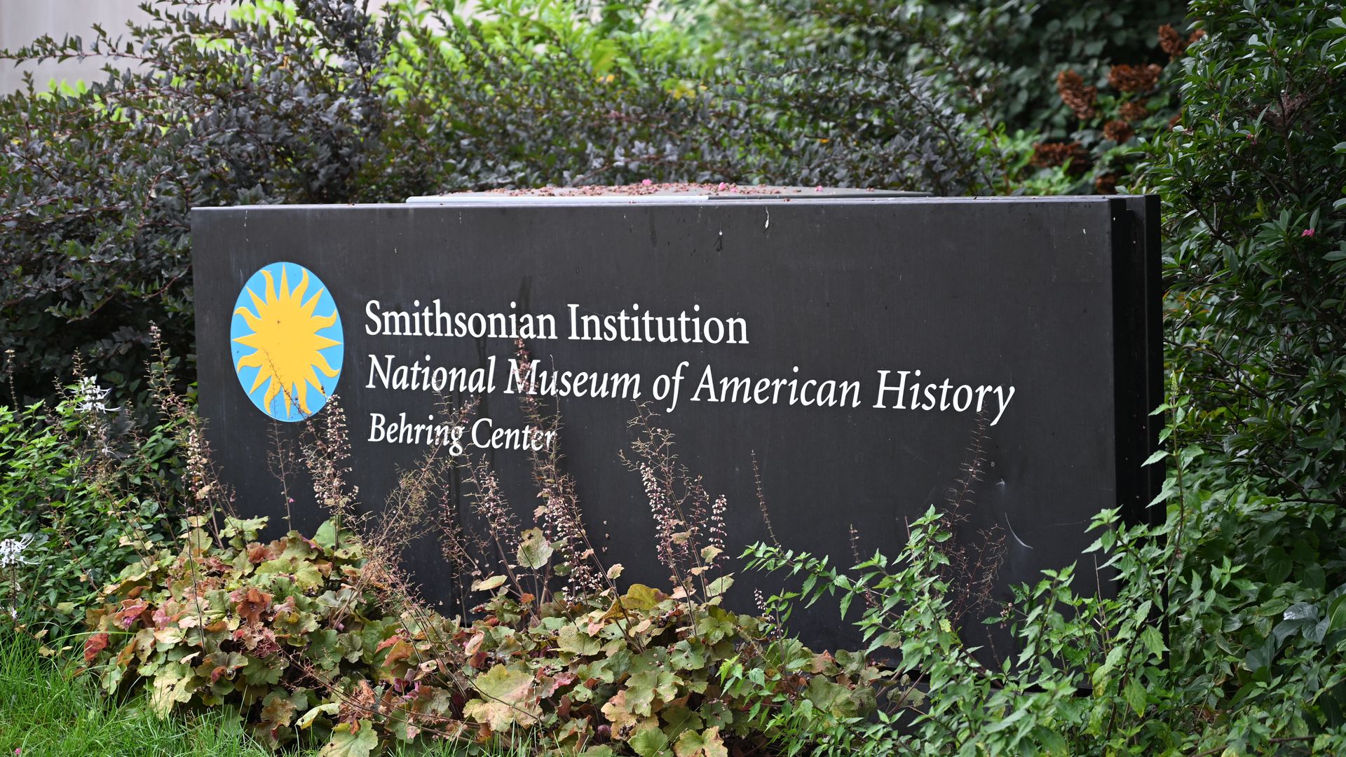 Black Smithsonian Institution sign with yellow sun logo, white text reading National Museum of American History Behring Center, surrounded by green and brown plants.