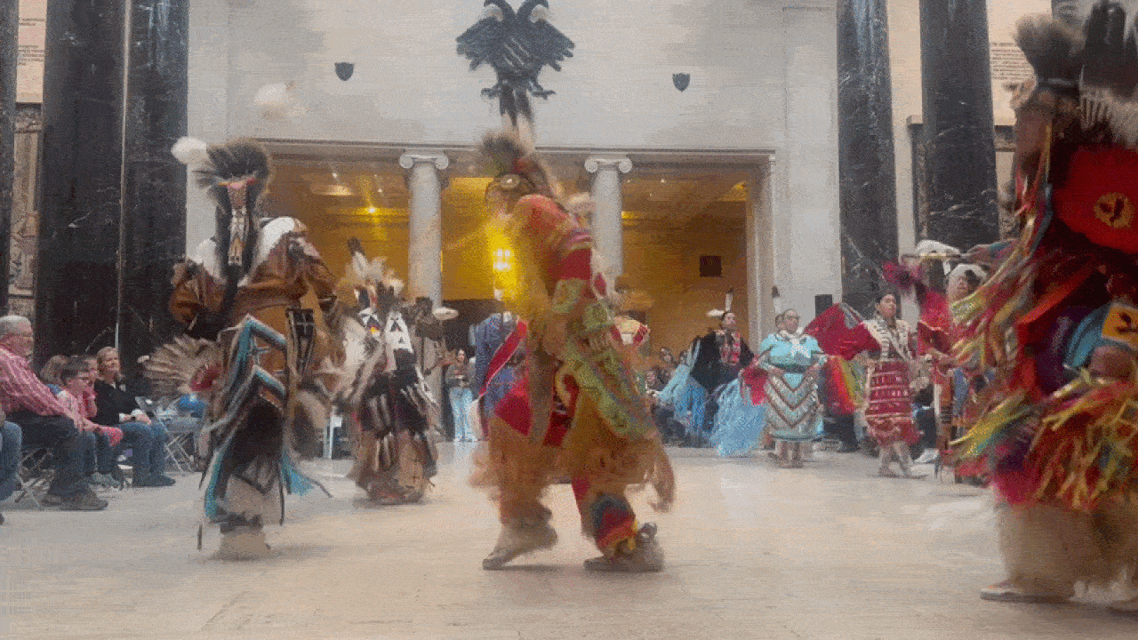 A group of dancers in colorful traditional Native American regalia performing in a large indoor space with columns and an eagle emblem above the entrance. Spectators watch nearby.