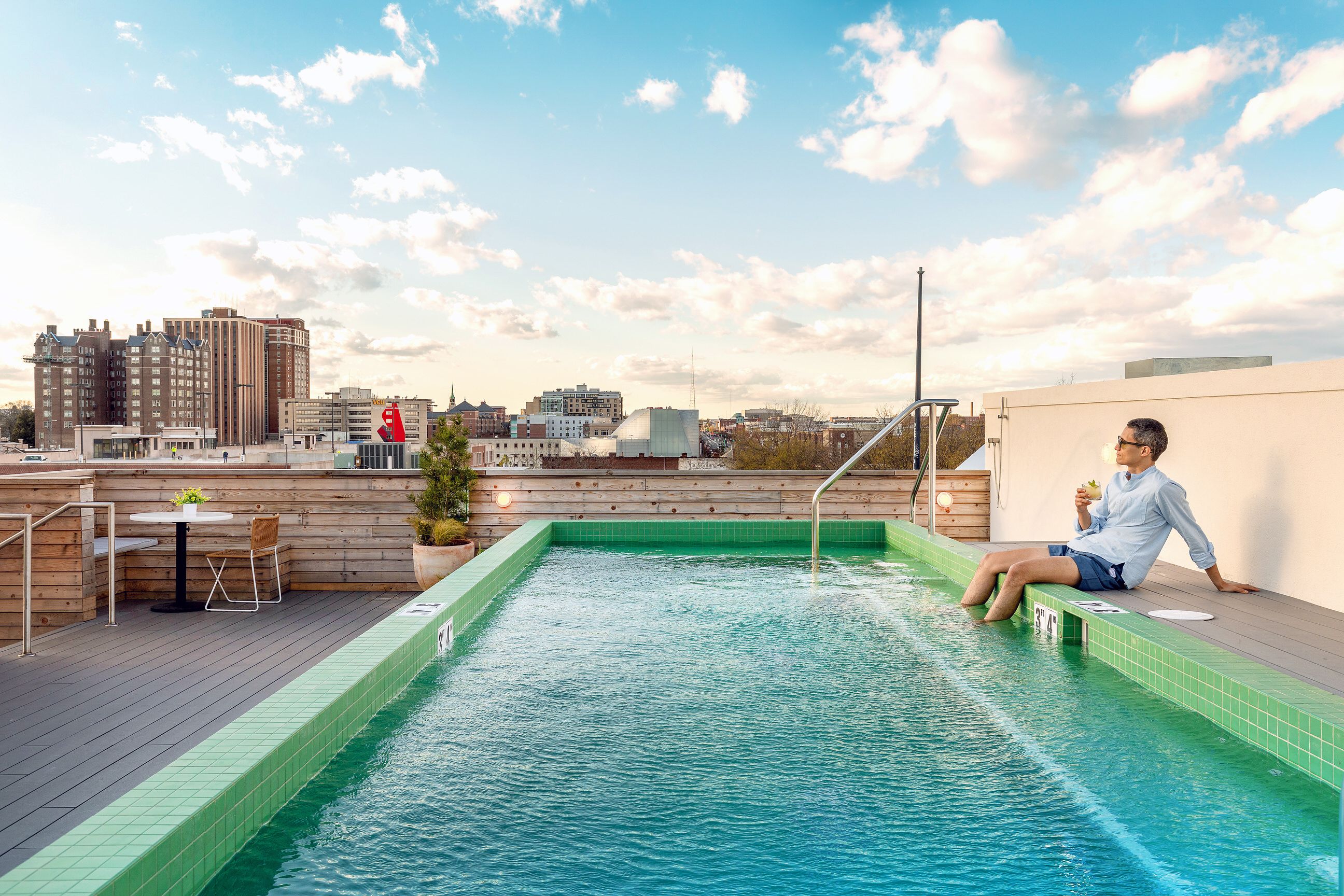 Rooftop swimming pool with a green-tiled edge and turquoise water; a man in a light blue shirt and shorts sits on the edge, sipping a drink, with a city skyline and blue sky behind.