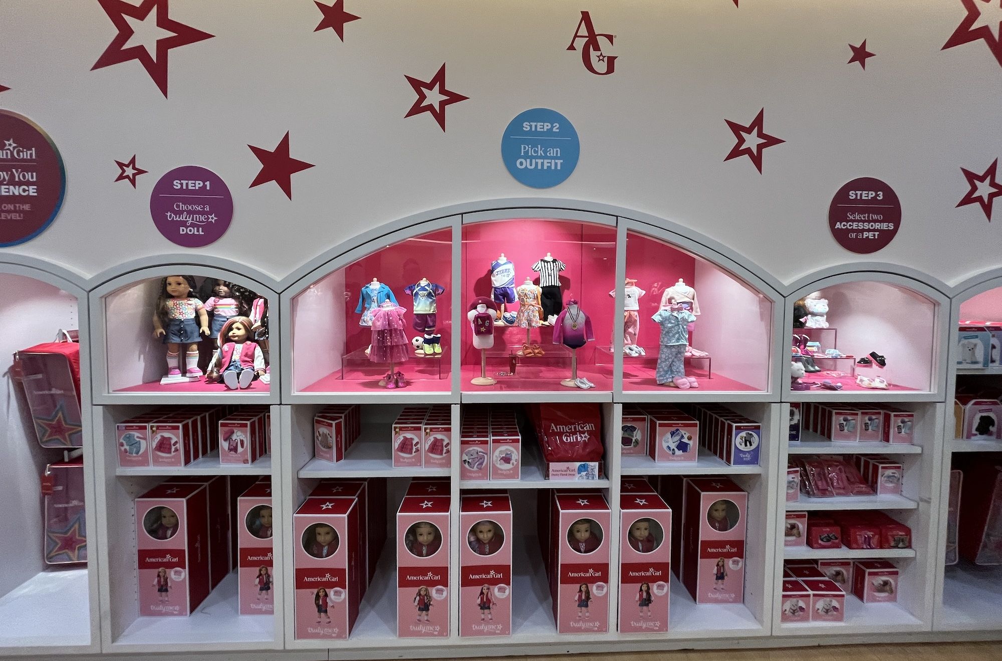 American Girl doll display with pink arched cases; dolls in outfits and boxed dolls on lower shelves; blue Step 2 sign "Pick an OUTFIT," purple Step 1 sign "Choose a truly me doll" on a white wall.