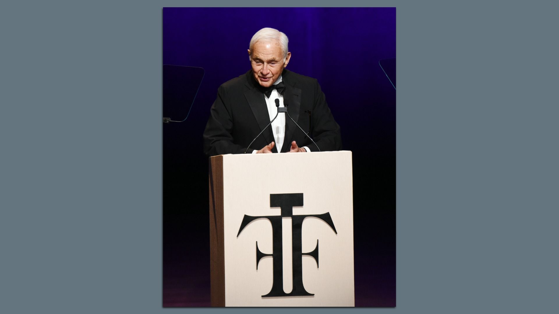 Elderly man in a tuxedo speaking at a podium with a large black 'TF' logo on a cream background, against a dark purple backdrop at an event.