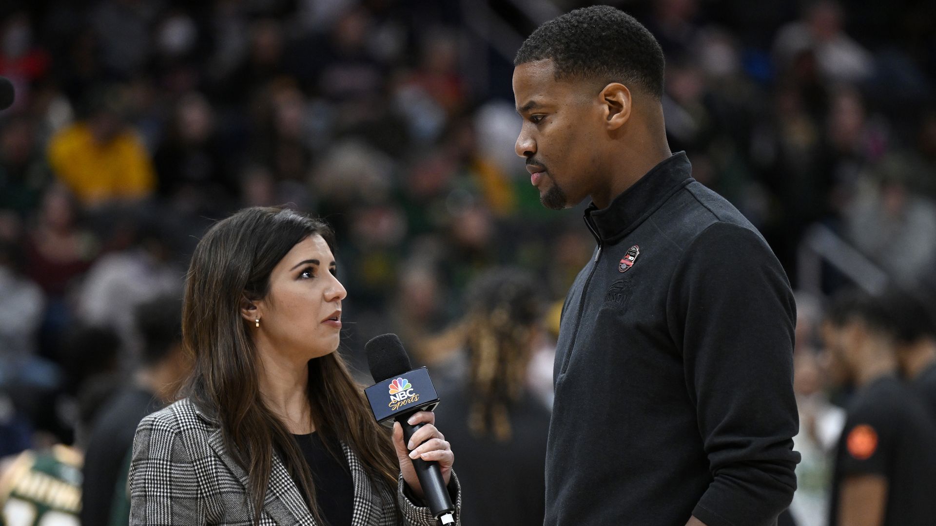 Photo of a reporter with a microphone interviewing a coach on the sidelines of a basketball game.