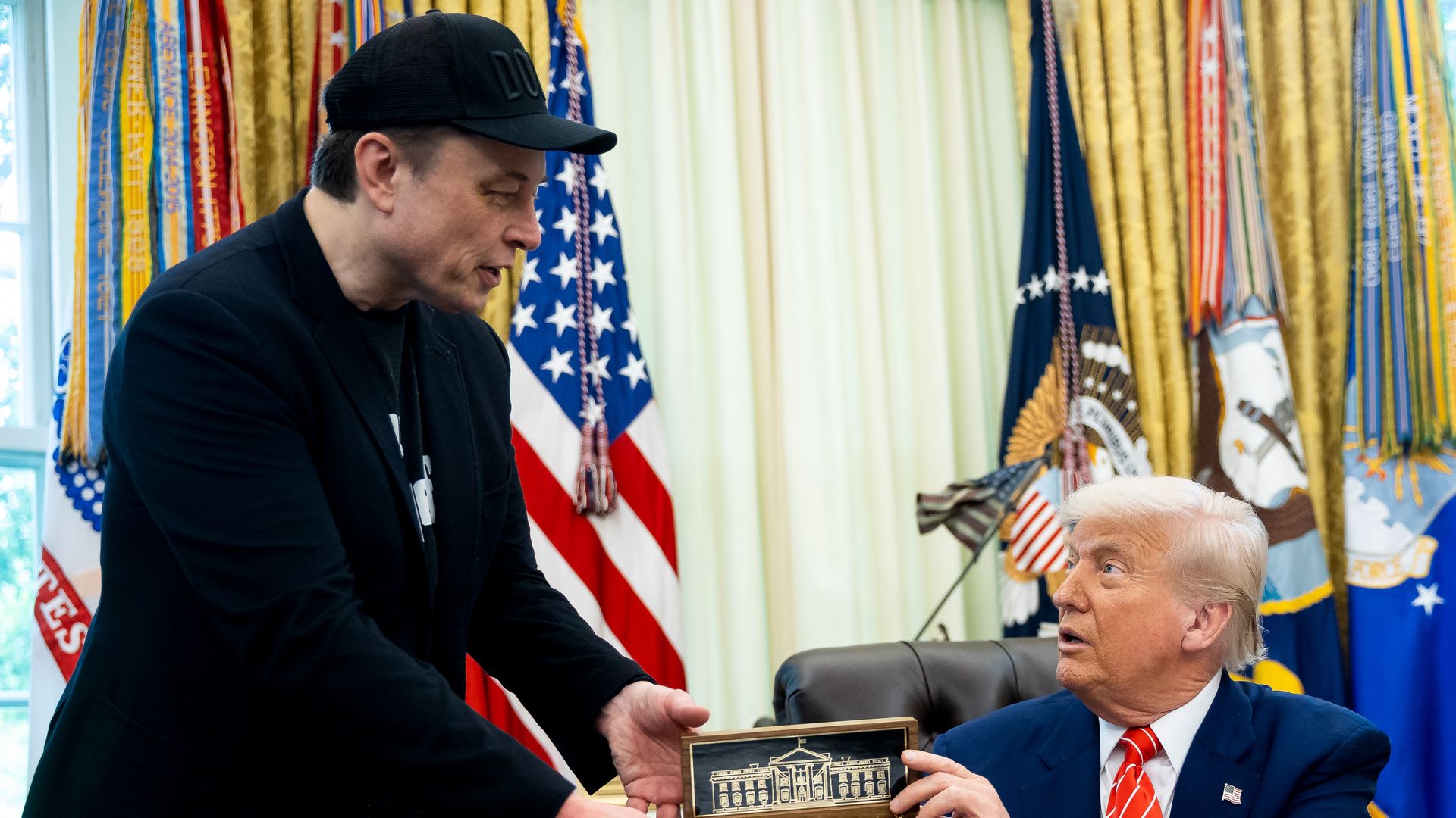 Donald Trump, wearing a blue suit, hands Elon Musk, wearing a black blazer and black hat, a key to the White House while sitting in a brown leather chair in front of gold and white curtains and a row of flags.
