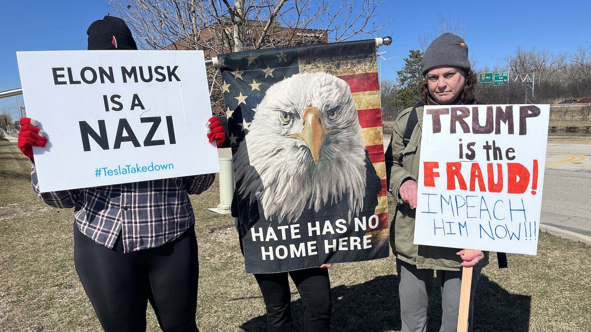 Three people holding protest signs, reading "Elon Musk is a Nazi," an American flag with an eagle reading "Hate Has No Home Here" and a third person wearing a knit cap holding a sign that reads "Trump is the Fraud! Impeach him now!!!"