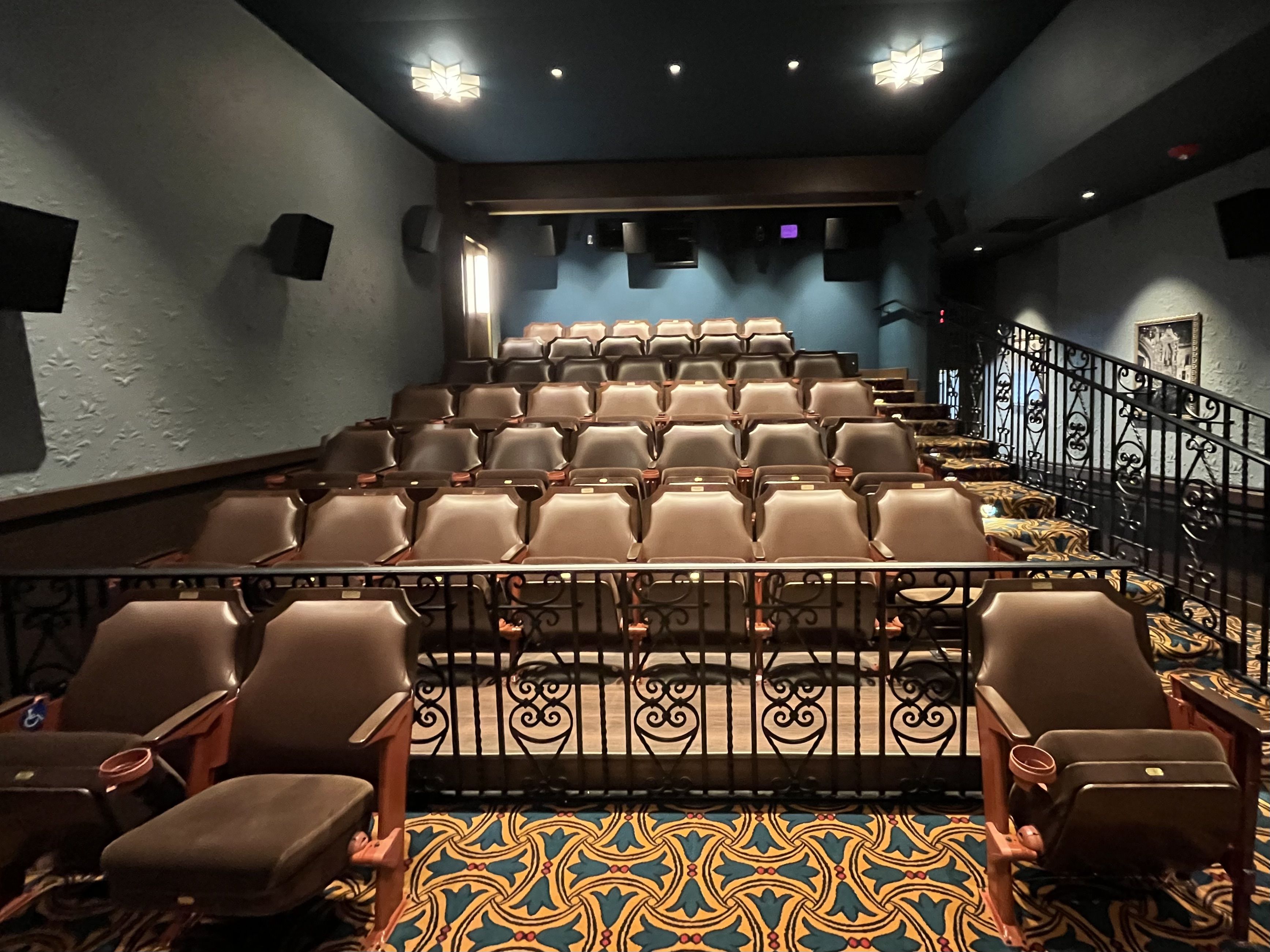 Small theater interior viewed from the front row: rows of tan seats, a black ornate railing in the foreground, teal-blue walls, a teal-and-gold patterned carpet, and a stage area at the back with lights.