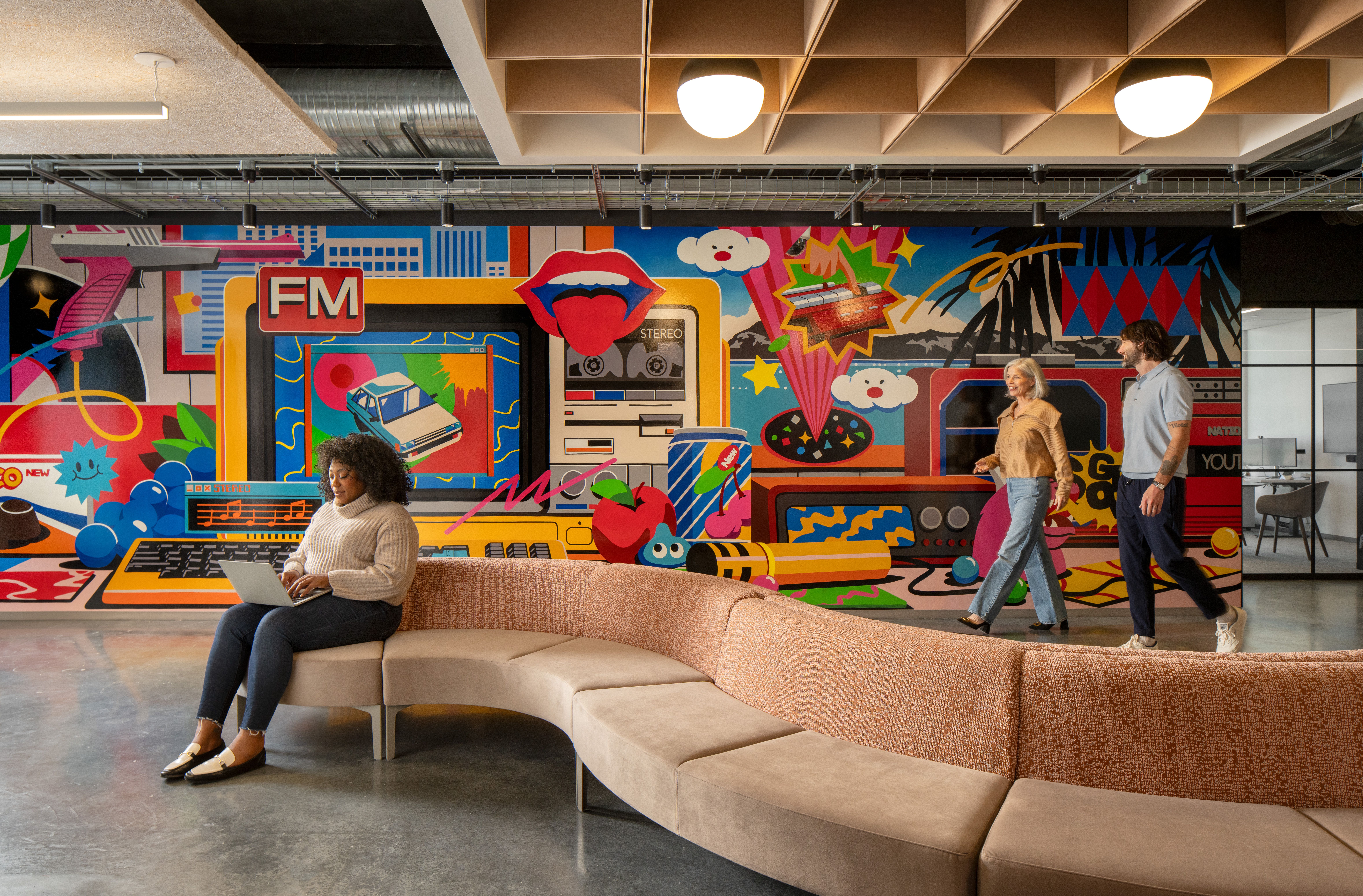 An employee types on a laptop while seating on a curved couch in front of a colorful mural 