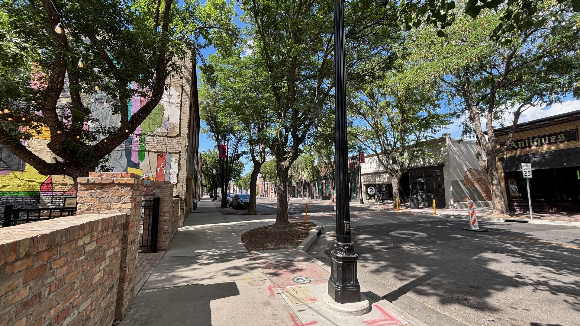 Sunlit street with tall trees. Left: colorful mural on a brick wall; string lights. Center: black lamppost. Right: shops, "Antiques" sign, and a parked car.