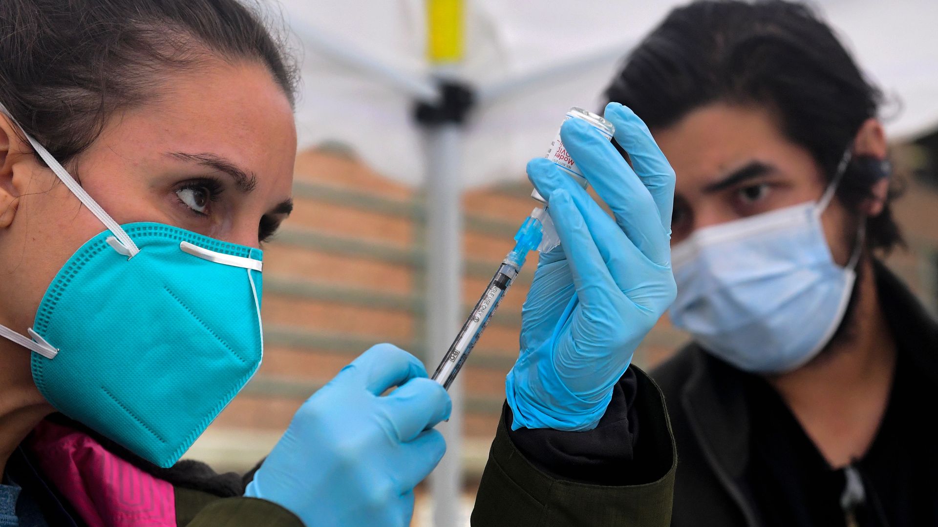 Photo of two masked people watching as one holds up a vaccine syringe