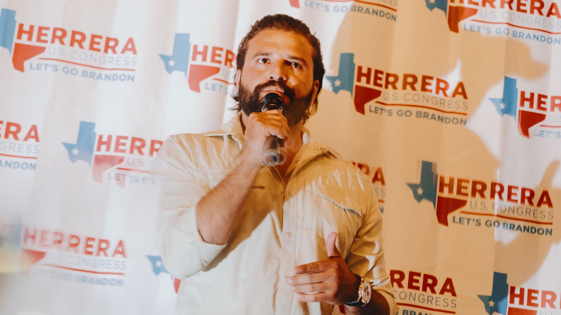 Republican congressional candidate Brandon Herrera — a man with brown hair and a beard wearing a neutral button-down shirt — speaks into a hand-held microphone in front of a wall covered in his campaign logo that reads 'HERRERA U.S. CONGRESS" and "LET'S GO BRANDON."