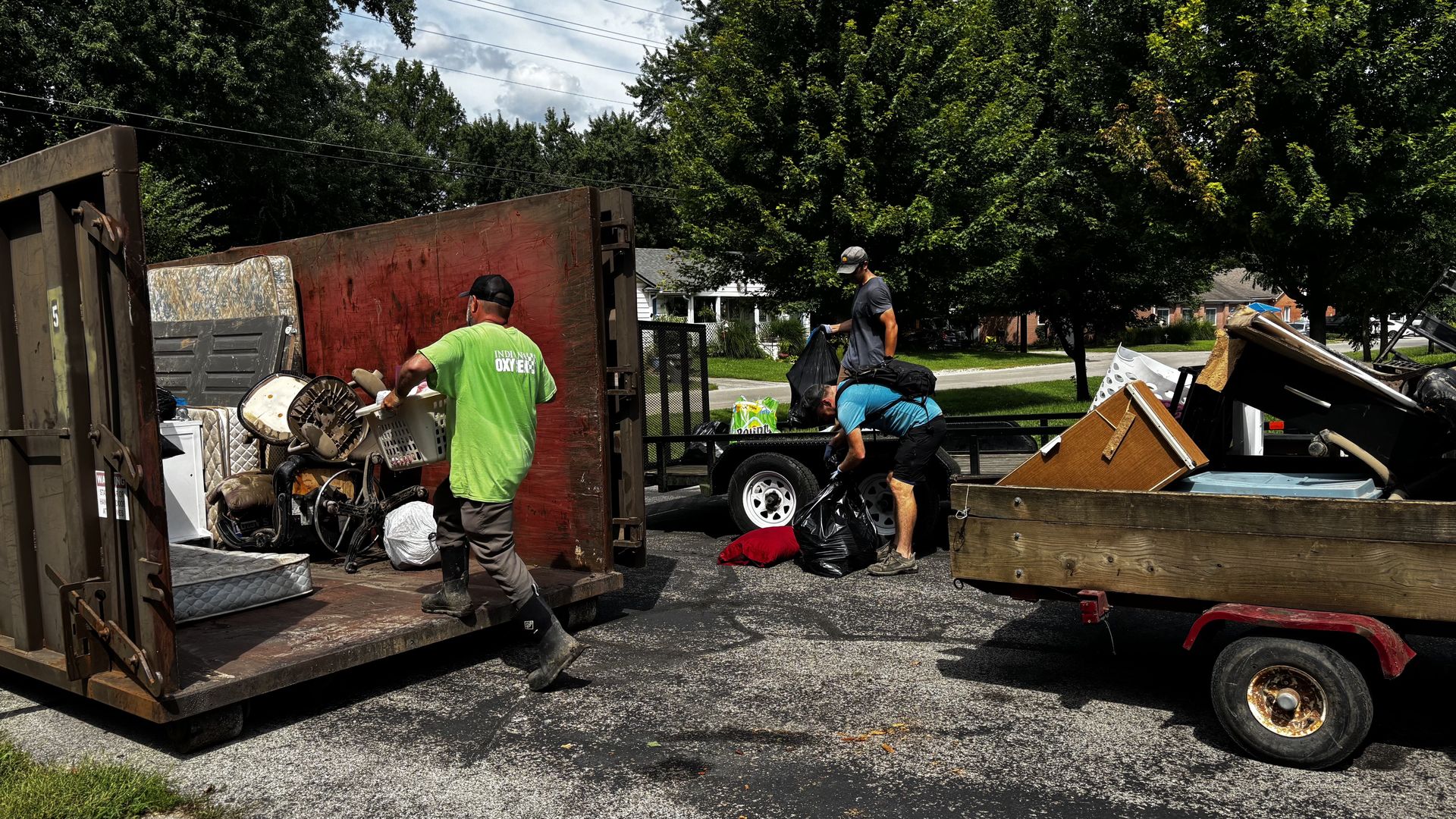 Three workers sort and unload trash from a rusted dumpster into a trailer on a residential street; furniture, bags, and appliances are visible, with green trees and houses behind.