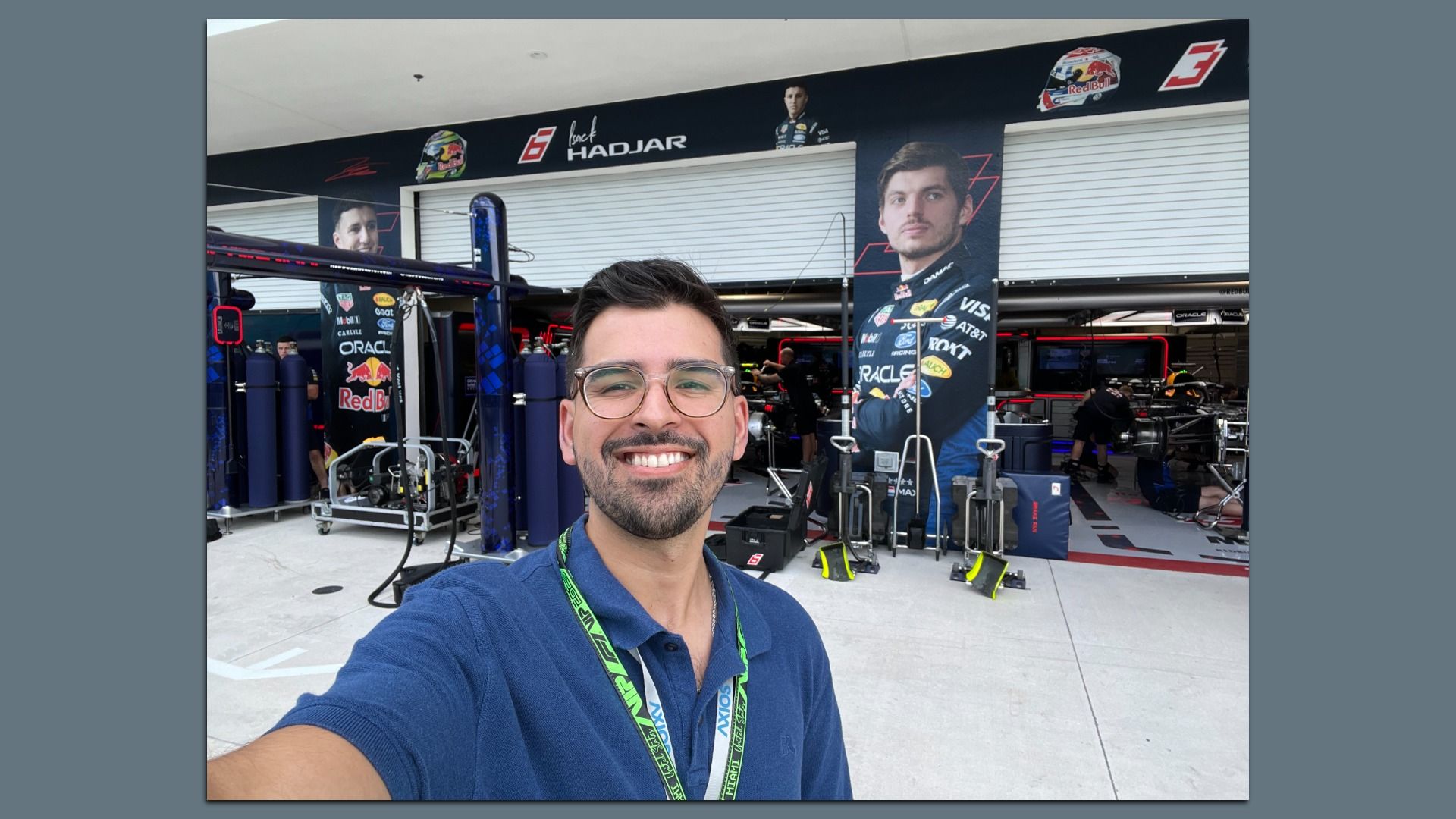Selfie of a smiling man with glasses wearing a blue polo and lanyard, taking a photo in a race team pit area with posters of drivers, Red Bull branding, and racing equipment in the background.