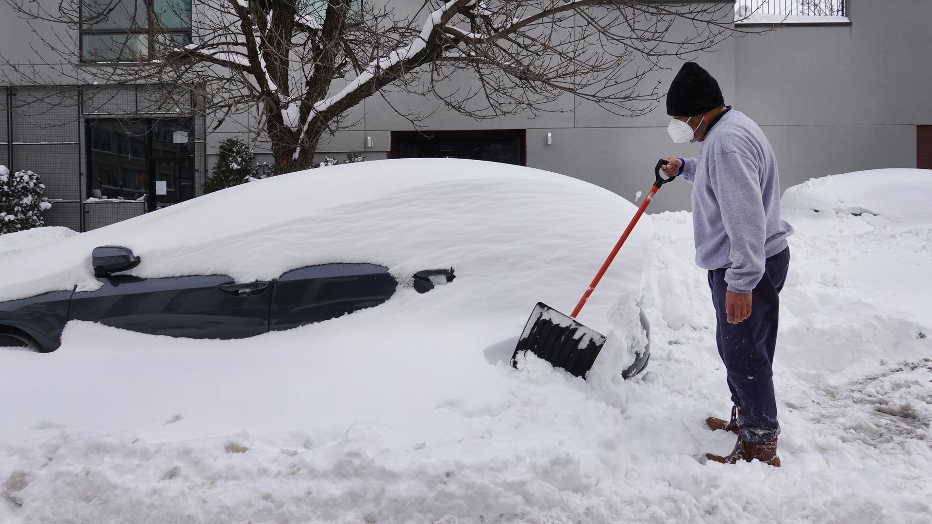 Picture of a man holding a shovel standing in front of a car buried in snow.