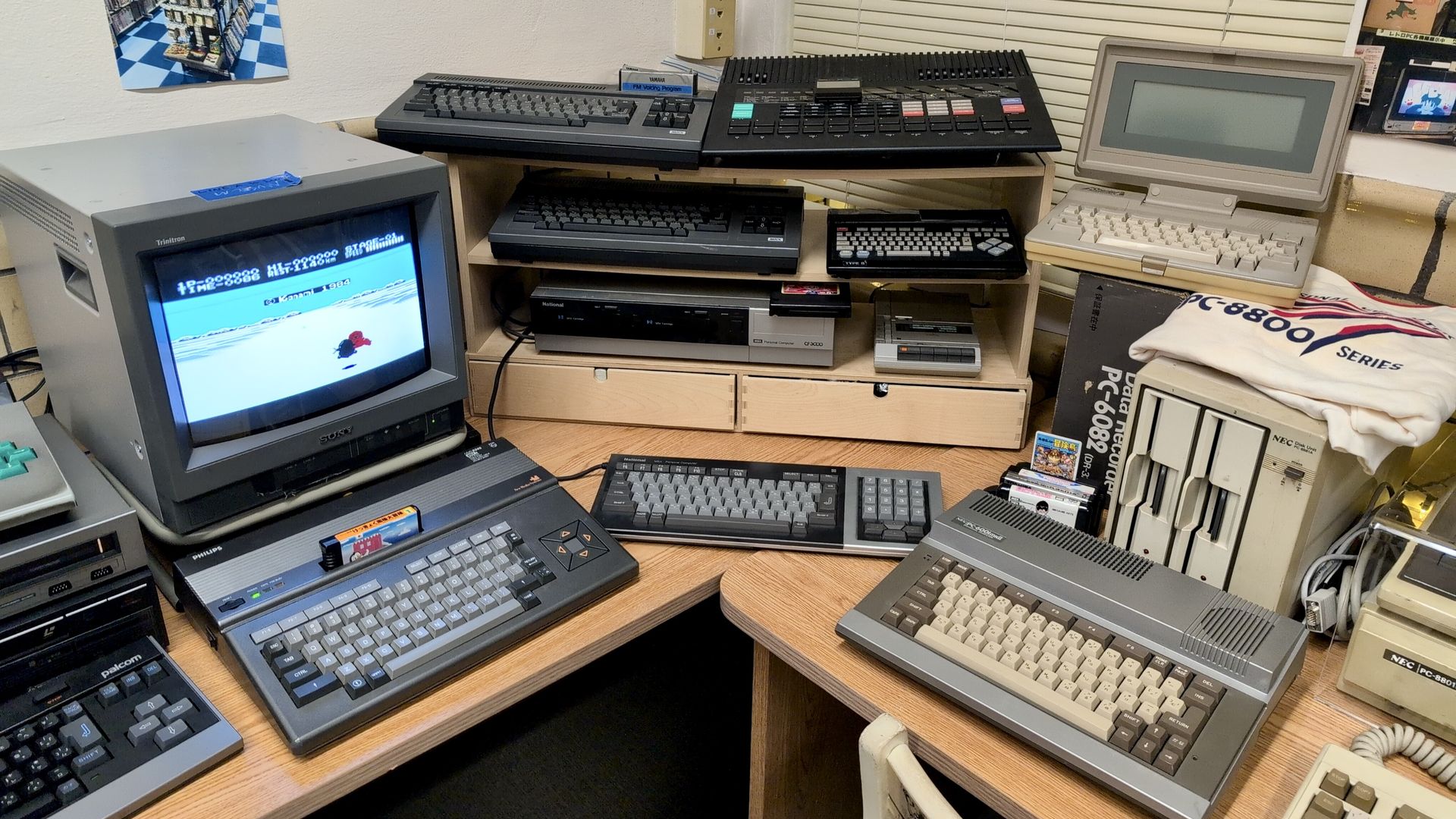 A collection of vintage computers and keyboards on desks and shelves, including a CRT monitor showing an 8-bit game, various keyboards, and computers with beige and gray color schemes.