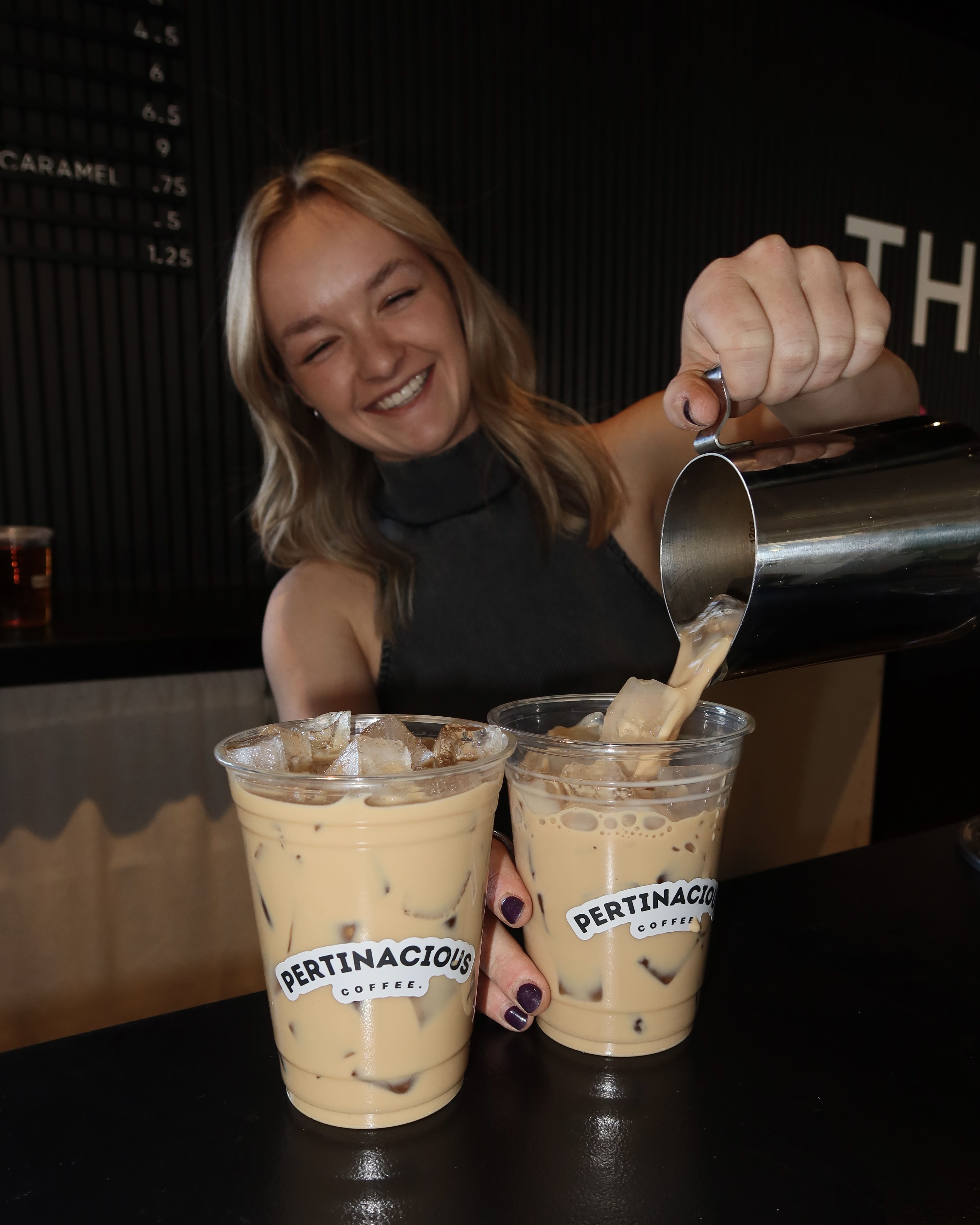 Smiling blonde woman pours iced coffee from a metal pitcher into two plastic cups labeled "PERTINACIOUS COFFEE" at a dark cafe counter.