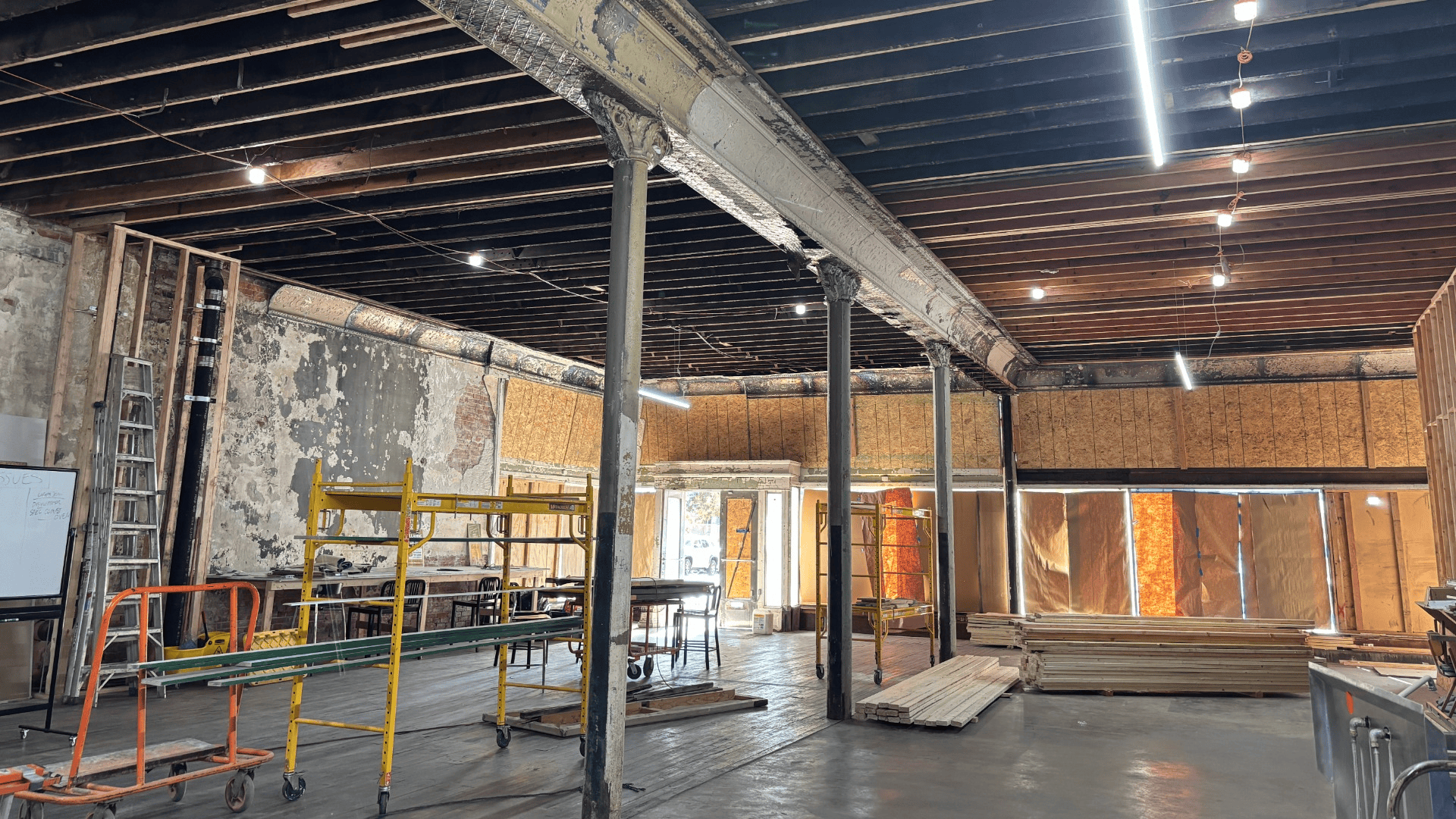 Interior of an unfinished renovation site with exposed ceiling joists and ductwork, tall metal columns, scaffolding, ladders, and stacked lumber against plywood walls.