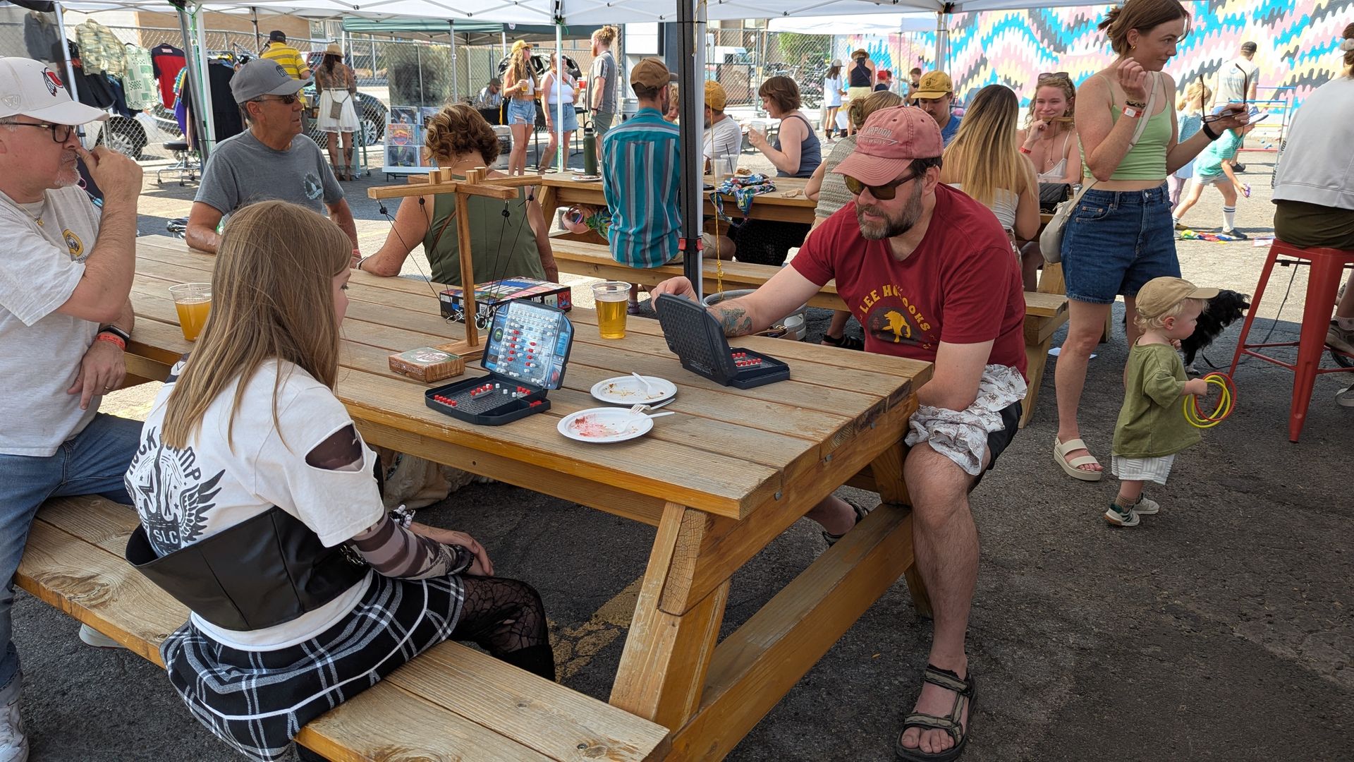 People sitting and standing around picnic tables under tents at an outdoor event, two playing a board game with red and white pieces, colorful mural on wall in background.
