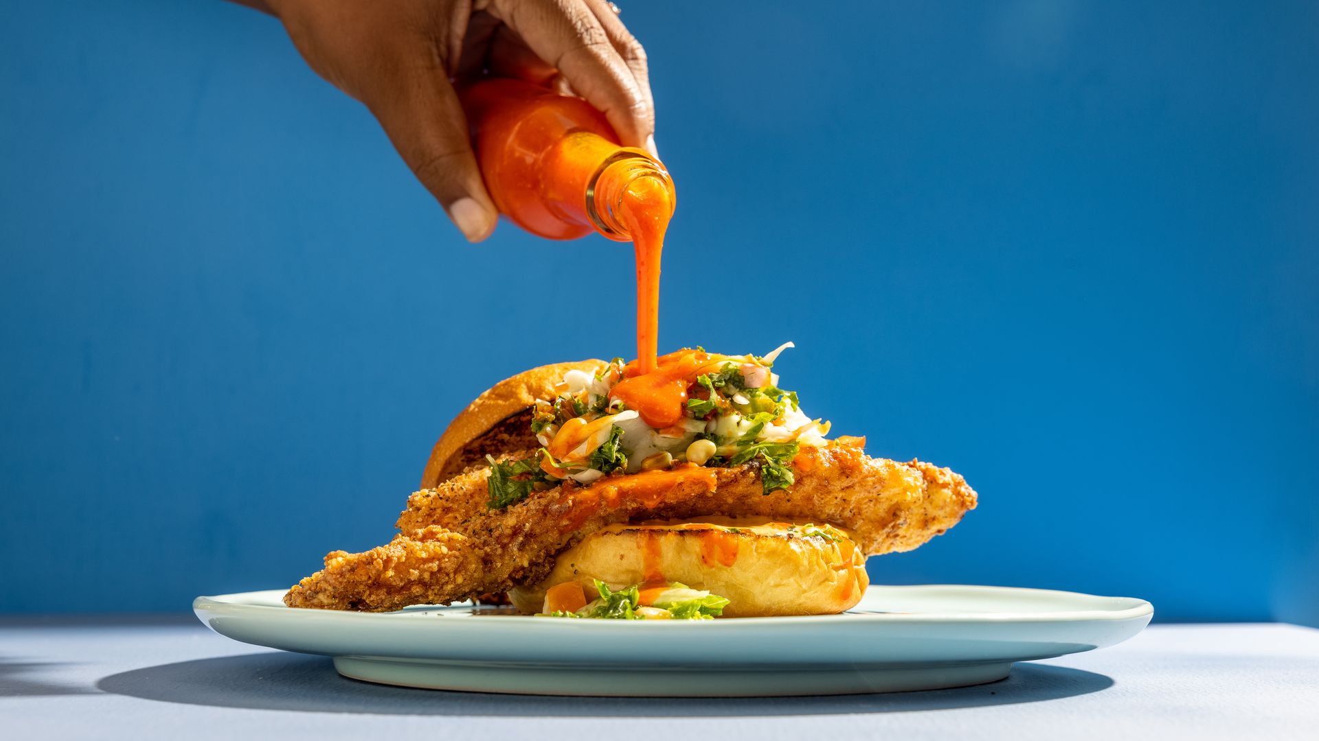 Hand pouring hot sauce over chicken sandwich
