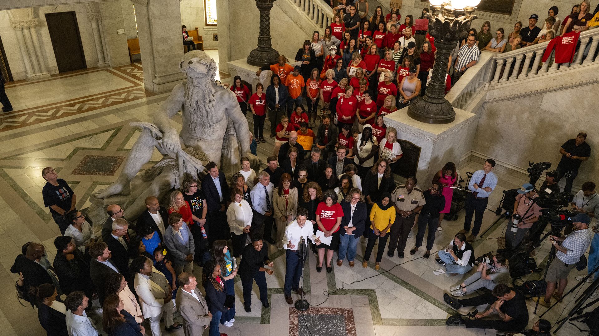 A large group of people gathered in a marble-floored hall with a large statue, some wearing red "Moms Demand Action" shirts, listening to a speaker at a microphone surrounded by media.