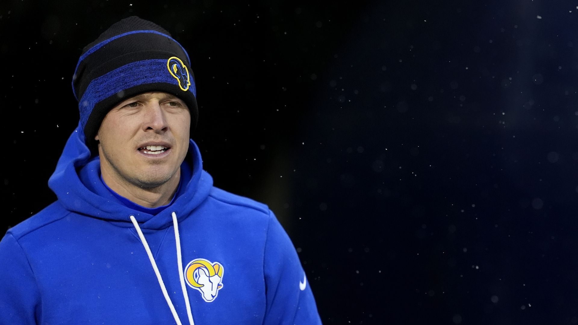 A man in a blue hooded sweatshirt with a white and yellow ram logo on the breast, and a blue and black striped wool cap with a blue ram logo on it, stares ahead against a black background. 