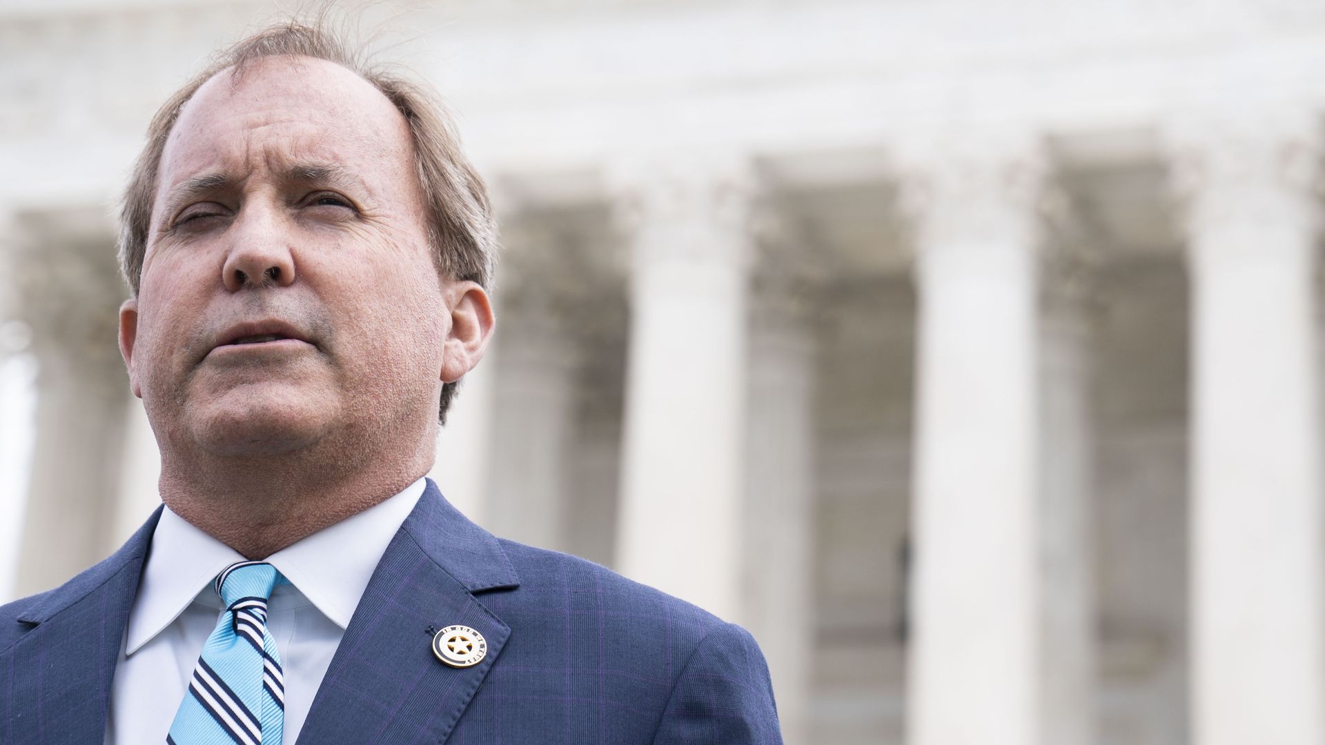 Ken Paxton, wearing a blue suit with a light blue tie, standing in front of a building with several Greek columns