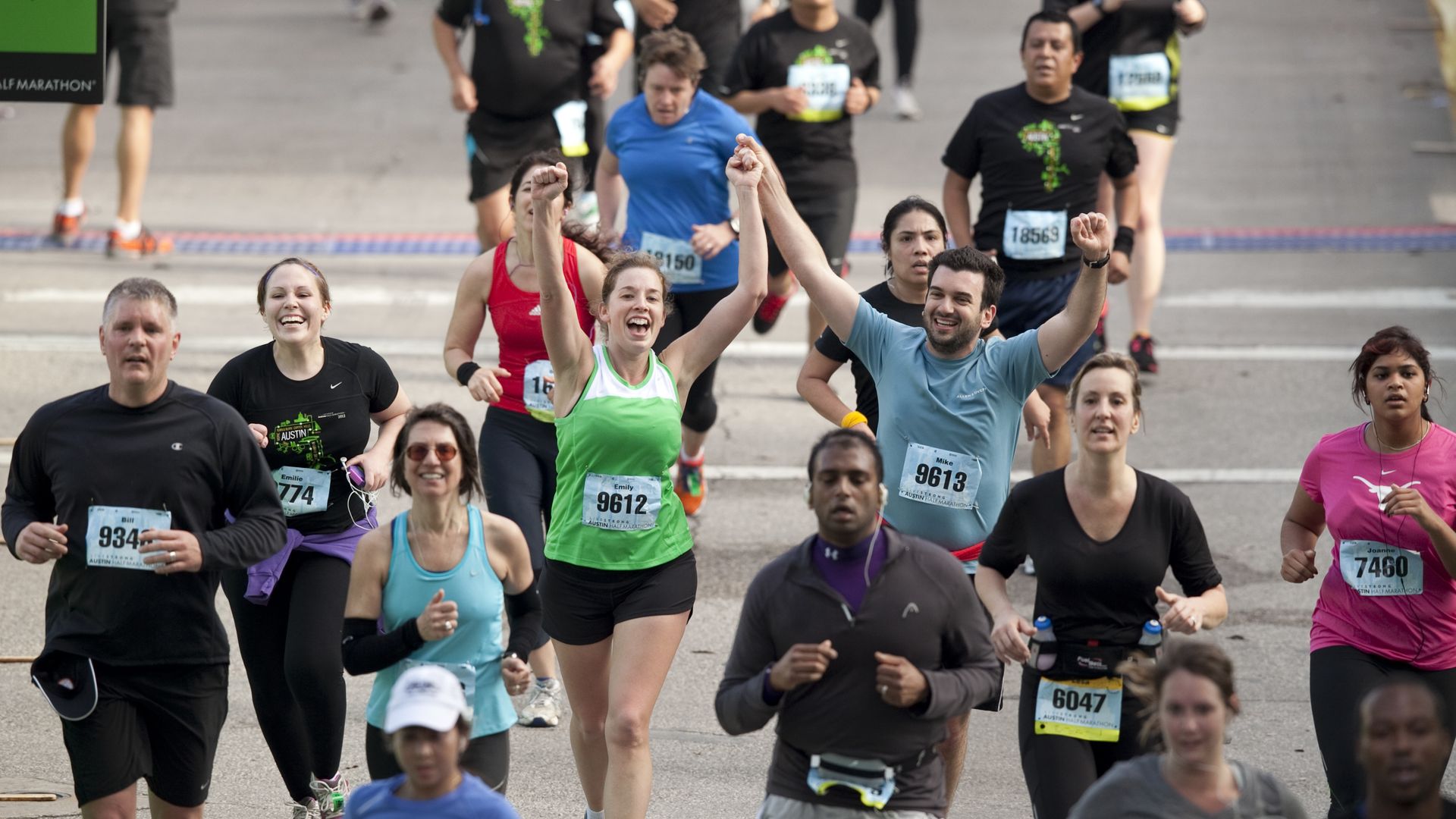 Runners of various ages and ethnicities participating in a marathon, some smiling and raising arms, wearing race bibs with numbers, on a city street with a race finish line visible.