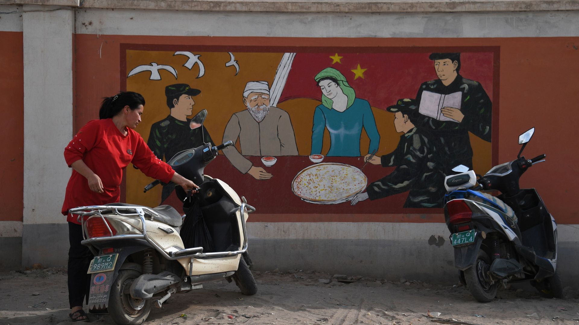 a Uighur woman beside a propaganda painting showing soldiers meeting with a Uighur family