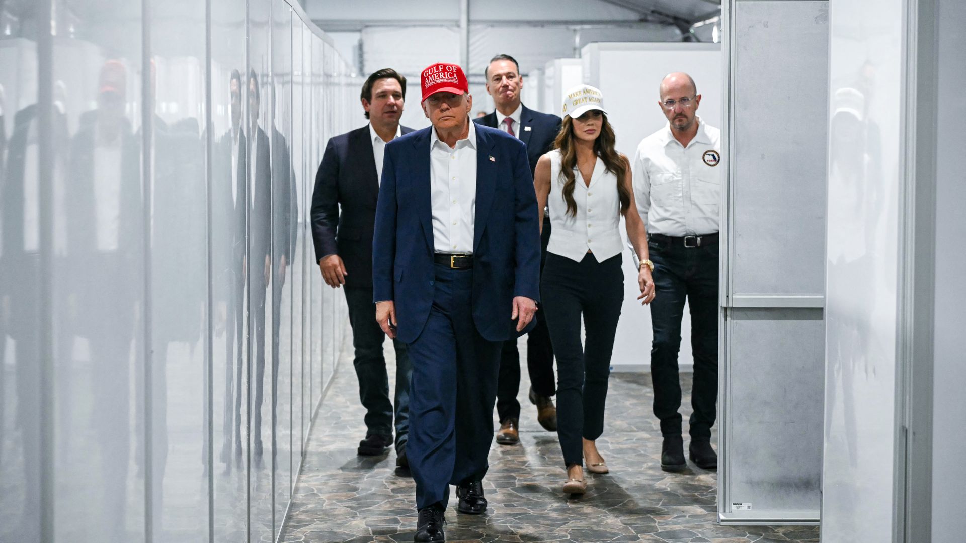 Five individuals walk down a hallway with white panel walls. The man in front wears a red 
