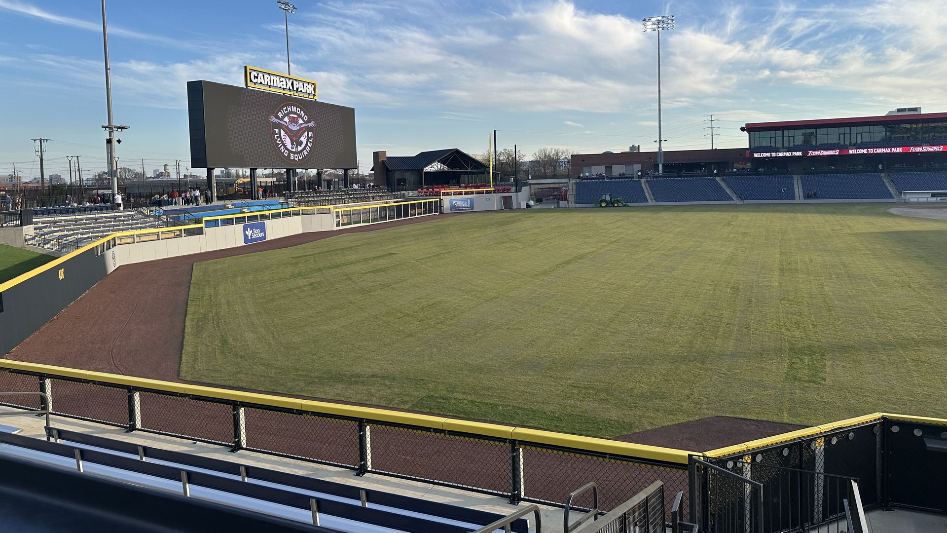 Wide view of a sunny baseball stadium with a large scoreboard reading "CARMAX PARK" and a team logo. Green infield and outfield, blue seats, yellow railing, empty stands, and tall light towers.