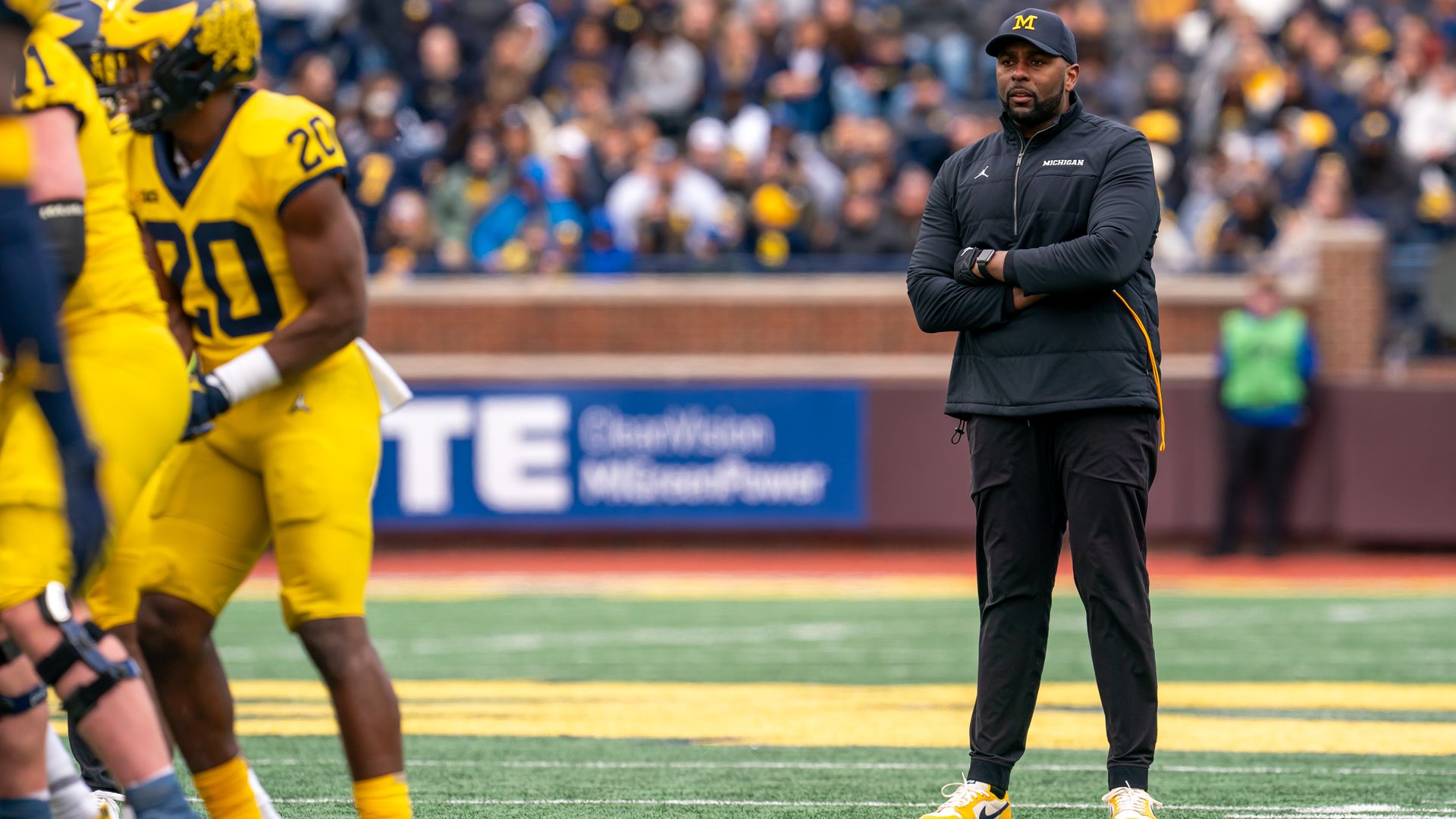 Michigan's coach stands with his arms folded on the field while players walk away. Crowd is in the background.
